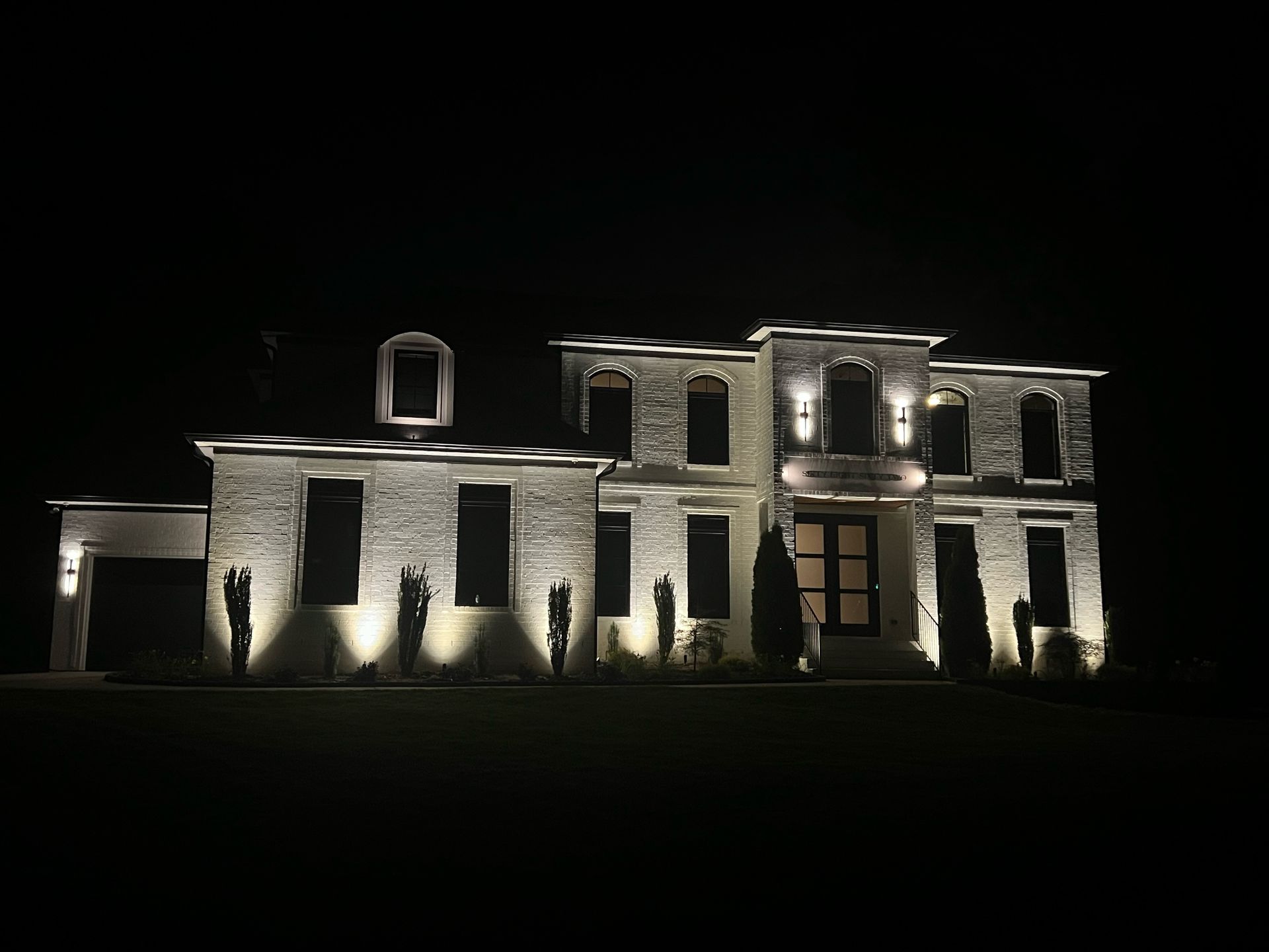 A large brick house illuminated at night with spotlights on the facade and landscaping. Dark sky surrounds the building.