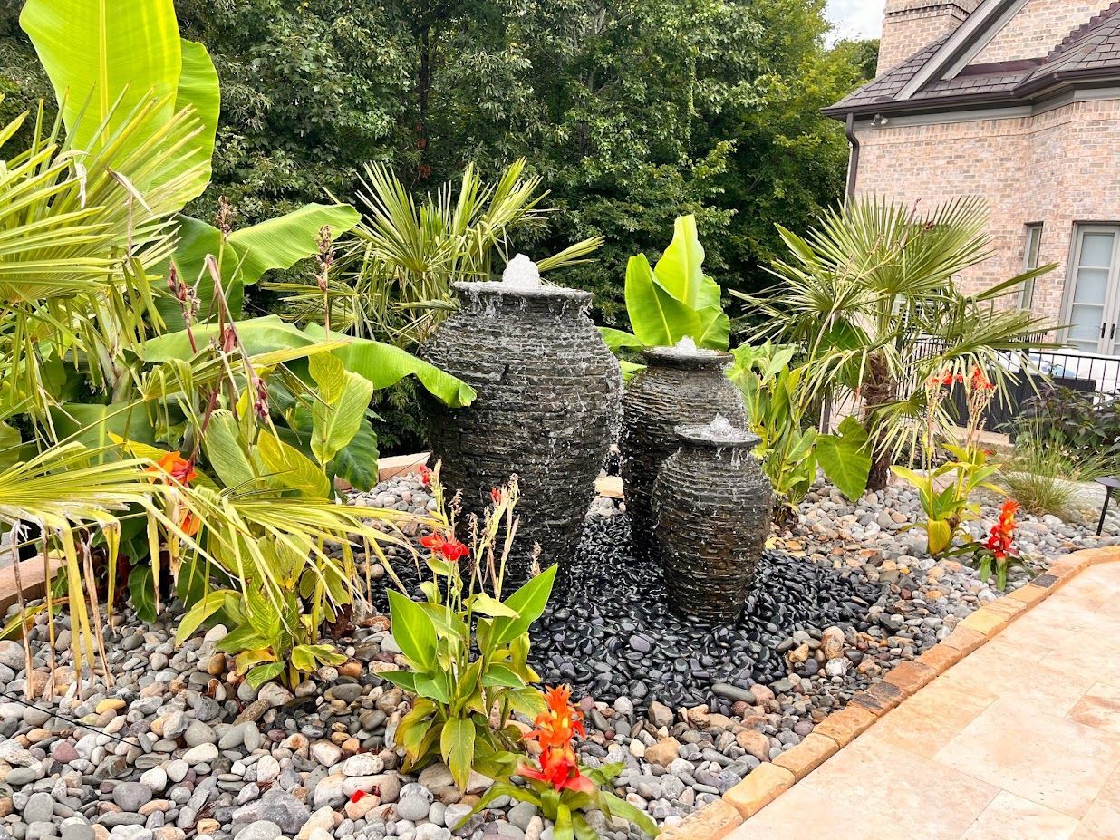 Stone water fountain with three urns in a garden with tropical plants and a brick patio.