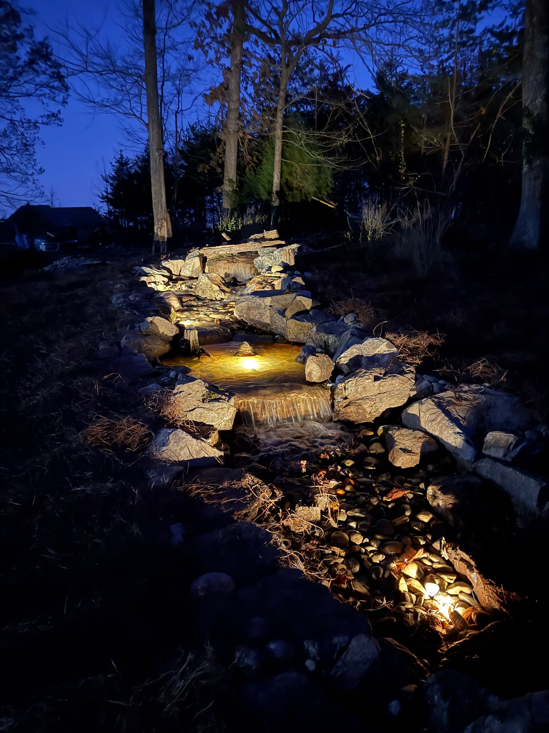 A lit waterfall cascading down rocks at night, illuminated by warm lights. Trees frame the background.