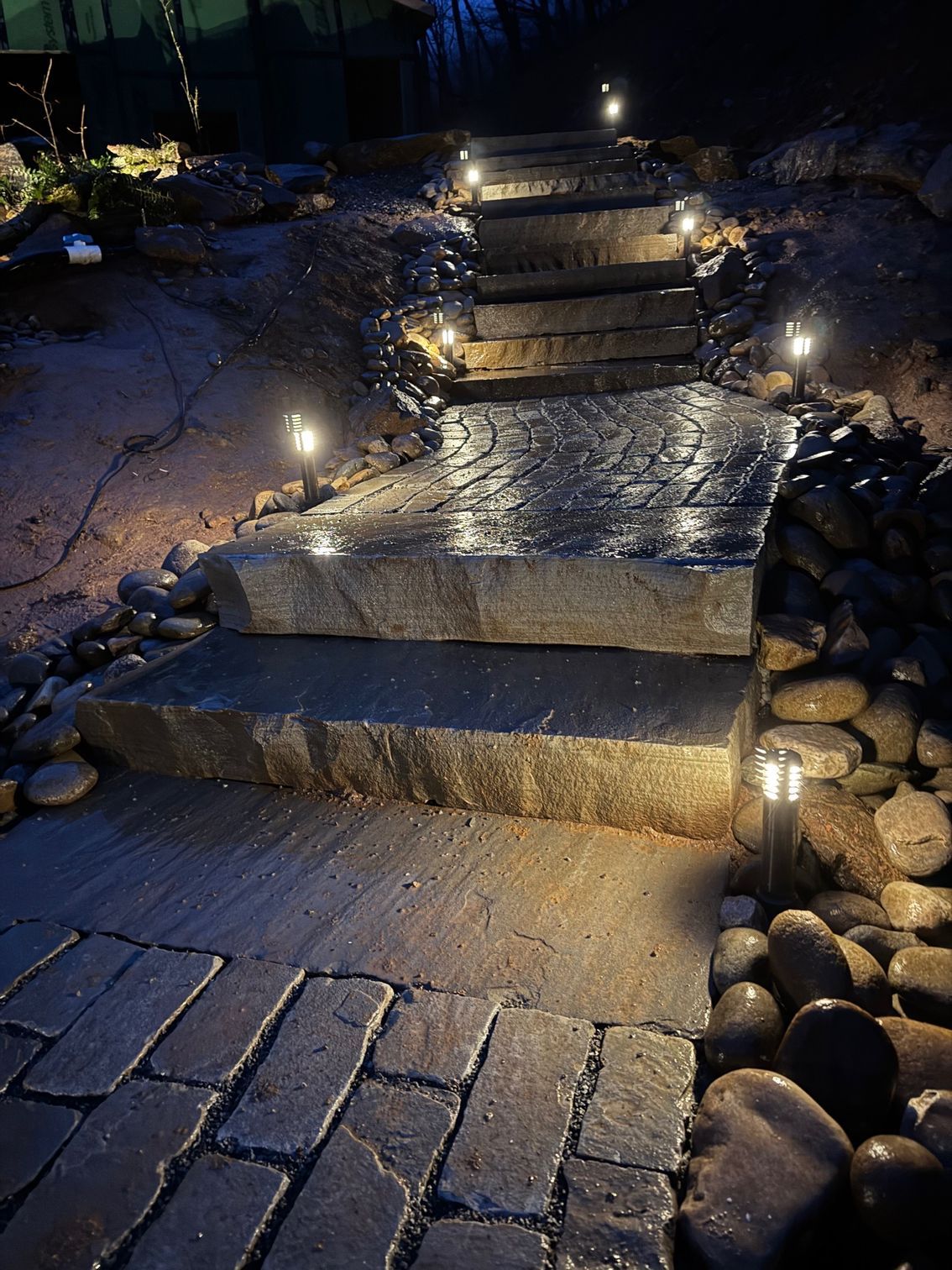 Stone steps with pathway lights leading uphill through a dark, outdoor setting. Wet cobblestones and rocks surround the steps.