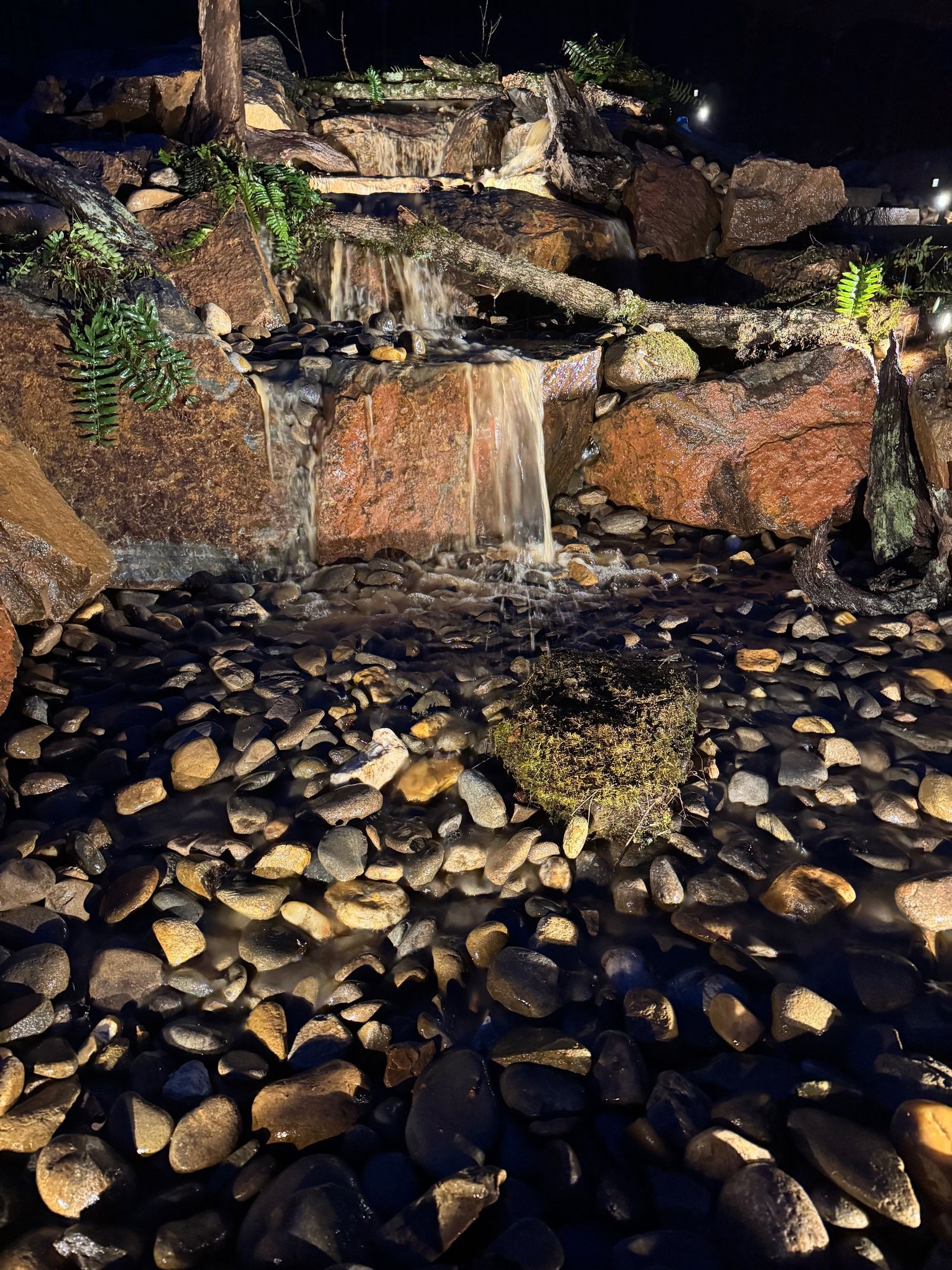 Waterfall cascading over brown rocks into a pool filled with stones, lit at night.