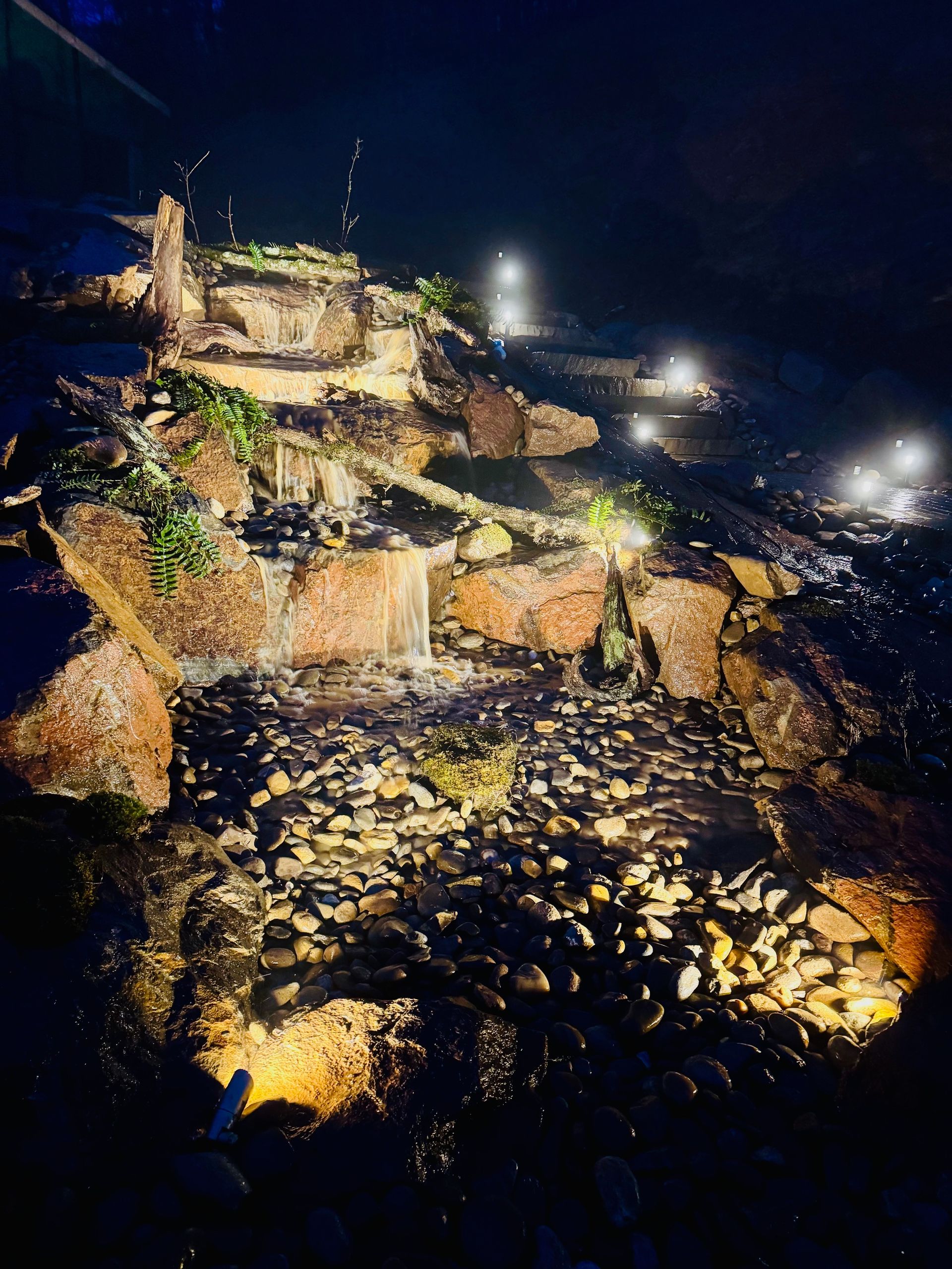 A nighttime view of a tiered waterfall illuminated by spotlights, cascading over rocky terrain with small shrubs.