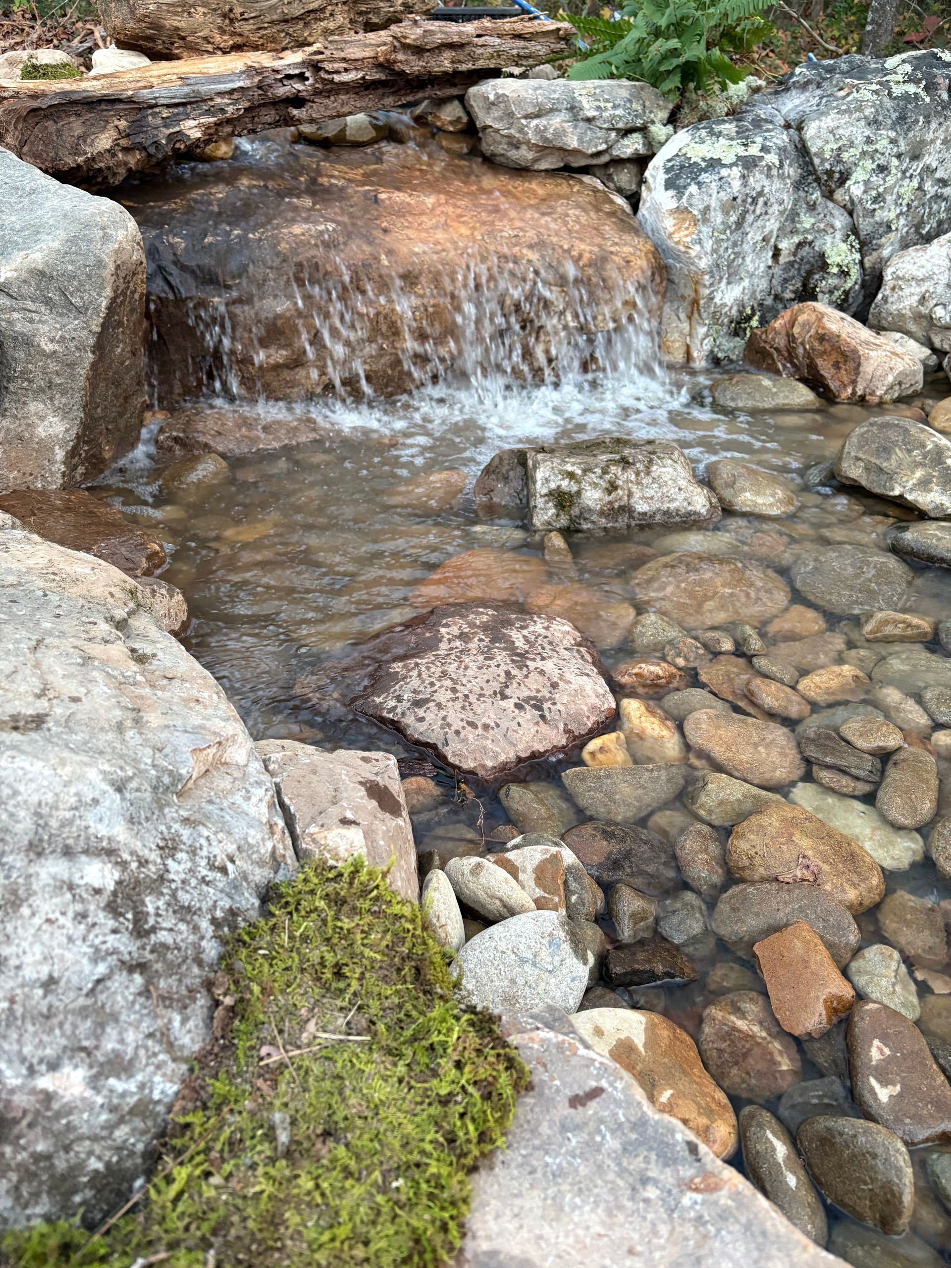 A small waterfall cascading over rocks into a stream bed filled with pebbles; moss grows on the edge.