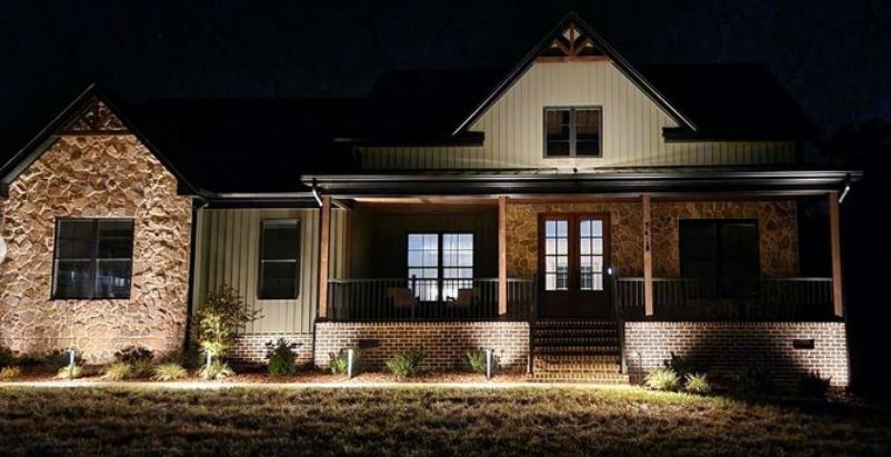A well-lit, stone-and-wood home at night, with lights highlighting the facade and surrounding landscaping.