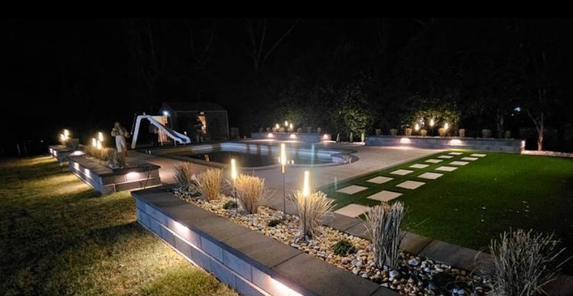 A nighttime view of a well-lit backyard with a pool and walkway. The scene is illuminated by warm-toned lights.