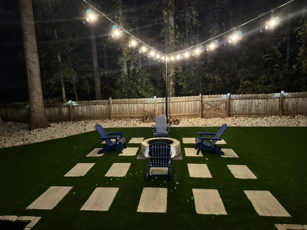 Nighttime backyard with string lights, a fire pit, blue Adirondack chairs, and stepping stones on a green lawn.