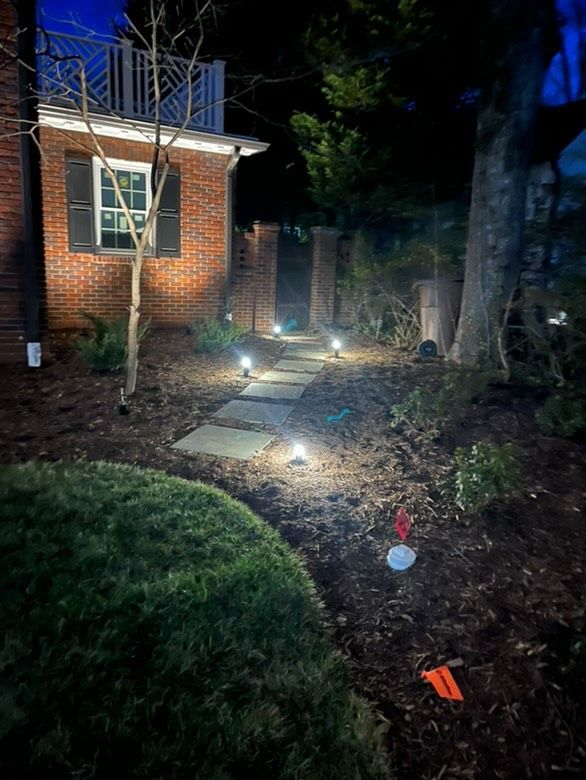 Pathway illuminated by ground lights leading to a brick wall in a night garden, with a house on the left.