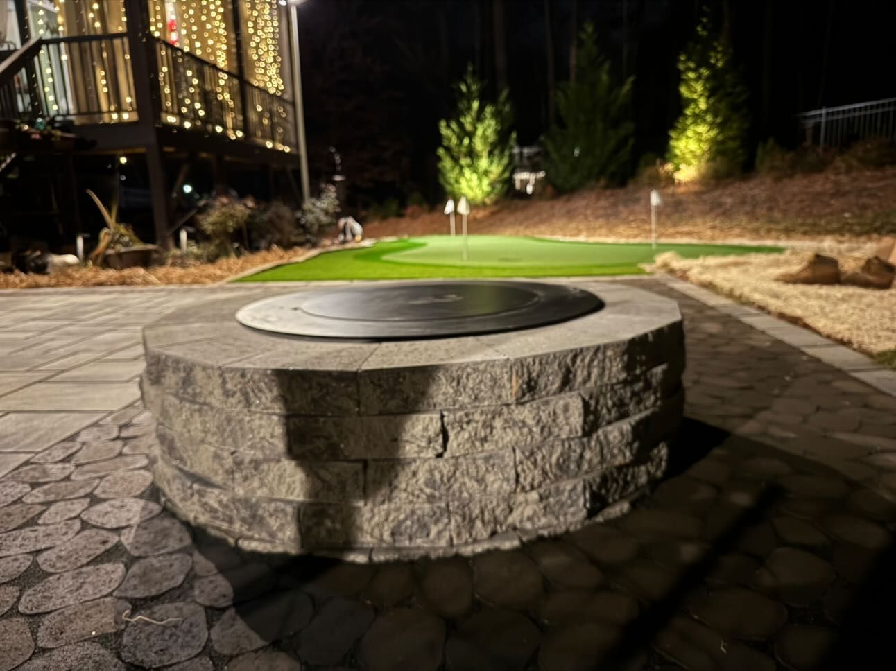 Circular stone fire pit with a black cover on a patio at night, golf green in background.