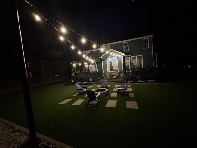 Night view of a backyard with string lights, chairs around a fire pit, and a two-story green house in the background.