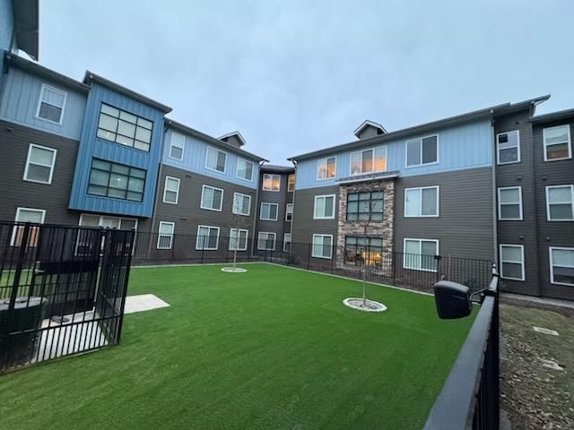 Exterior view of a modern apartment complex with a central green courtyard. The building has blue and gray siding with multiple windows.