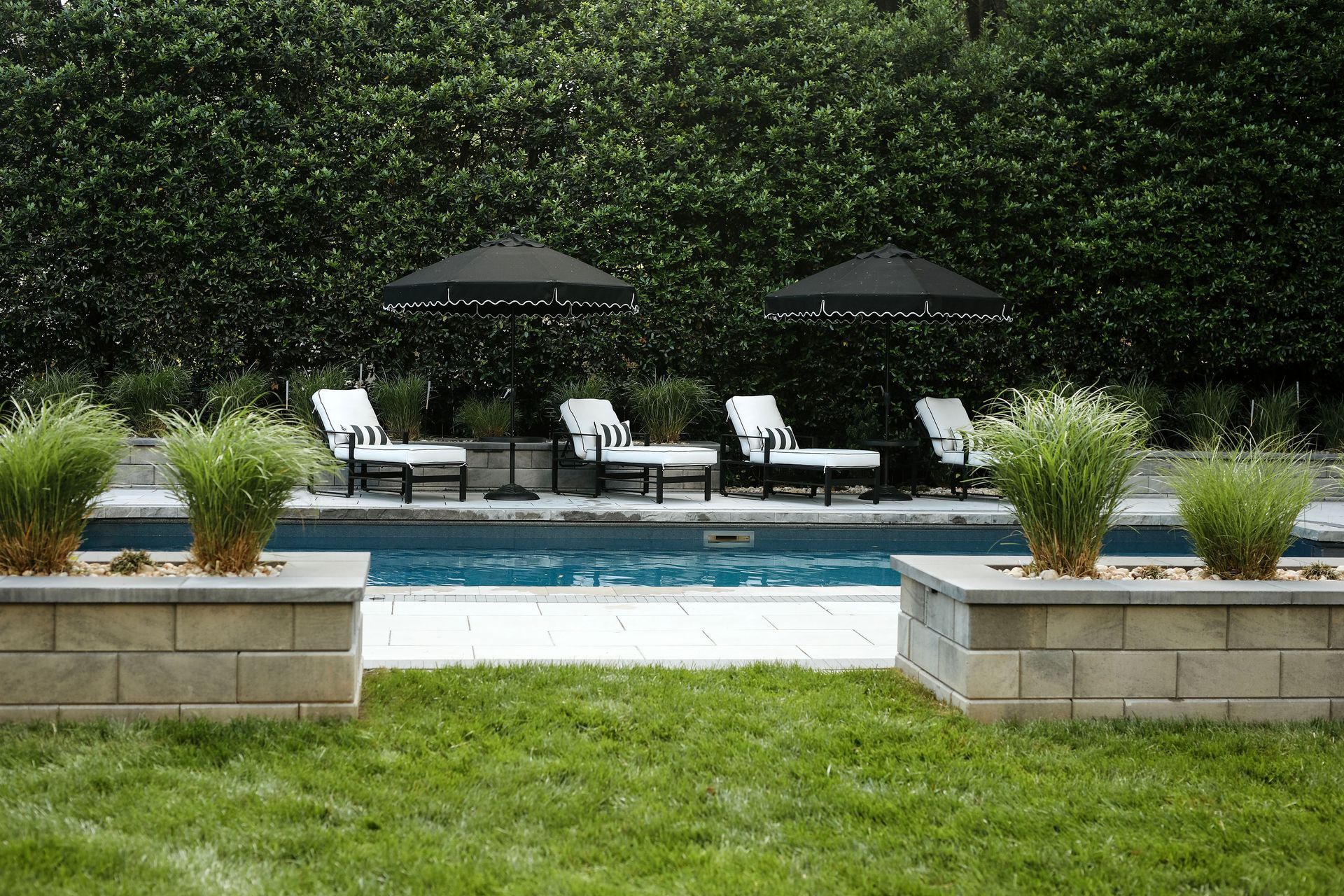 A backyard pool with lounge chairs under black umbrellas, backed by a green hedge. Lawn and stone planters are in the foreground.