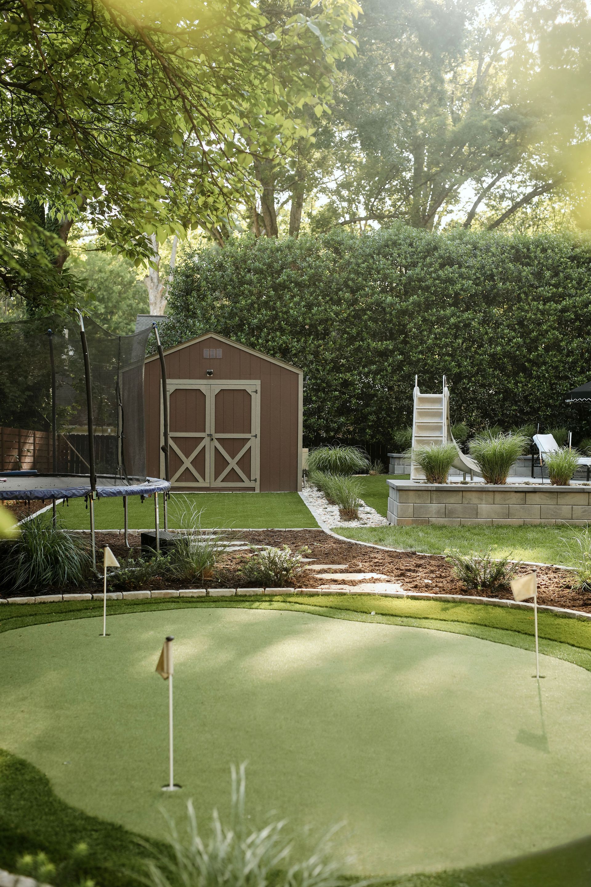 Backyard scene with a putting green in the foreground, shed and trampoline in the background, lush greenery and landscaping.