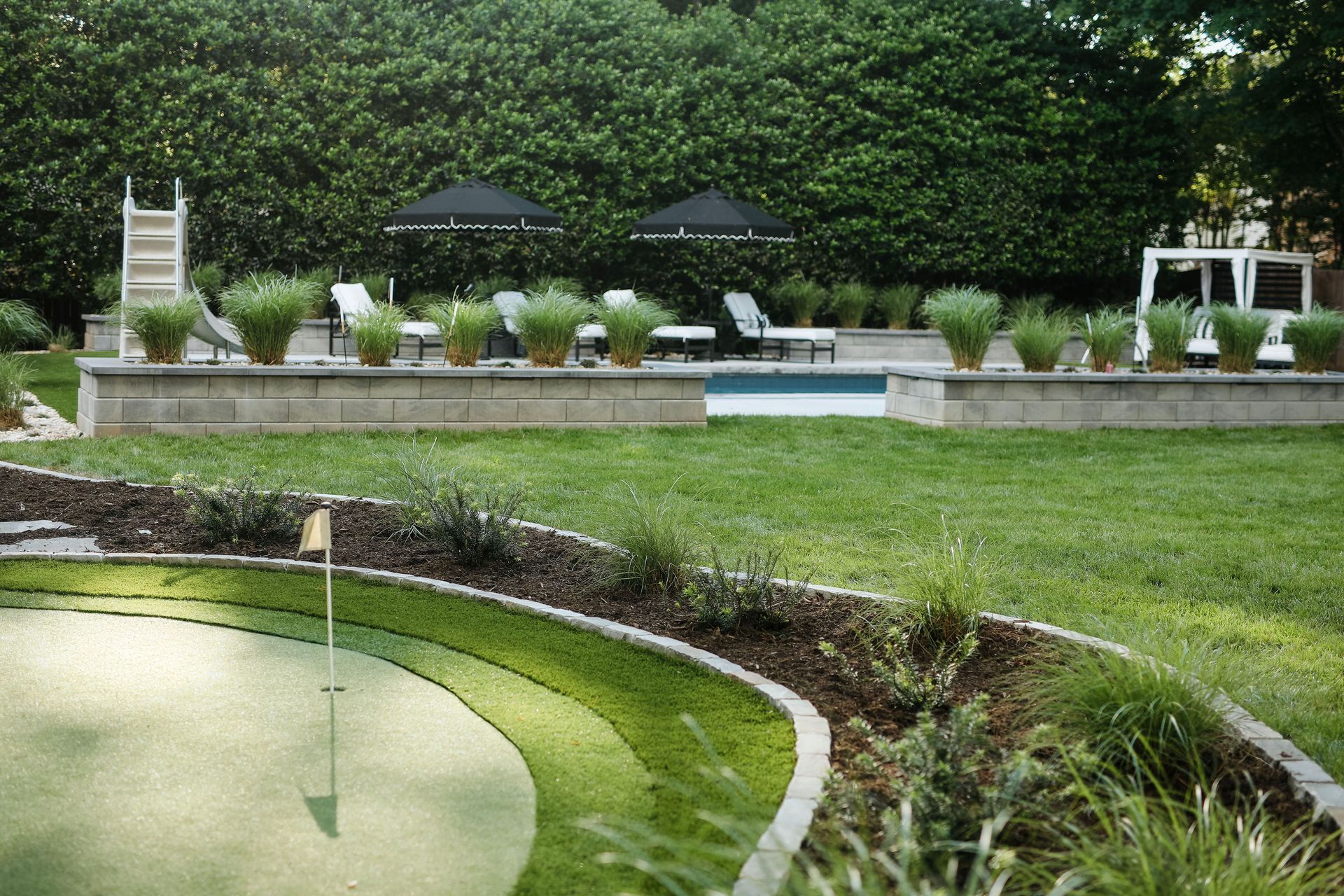 Green putting green in foreground, backyard with pool and lounge chairs, lush hedges in the background.