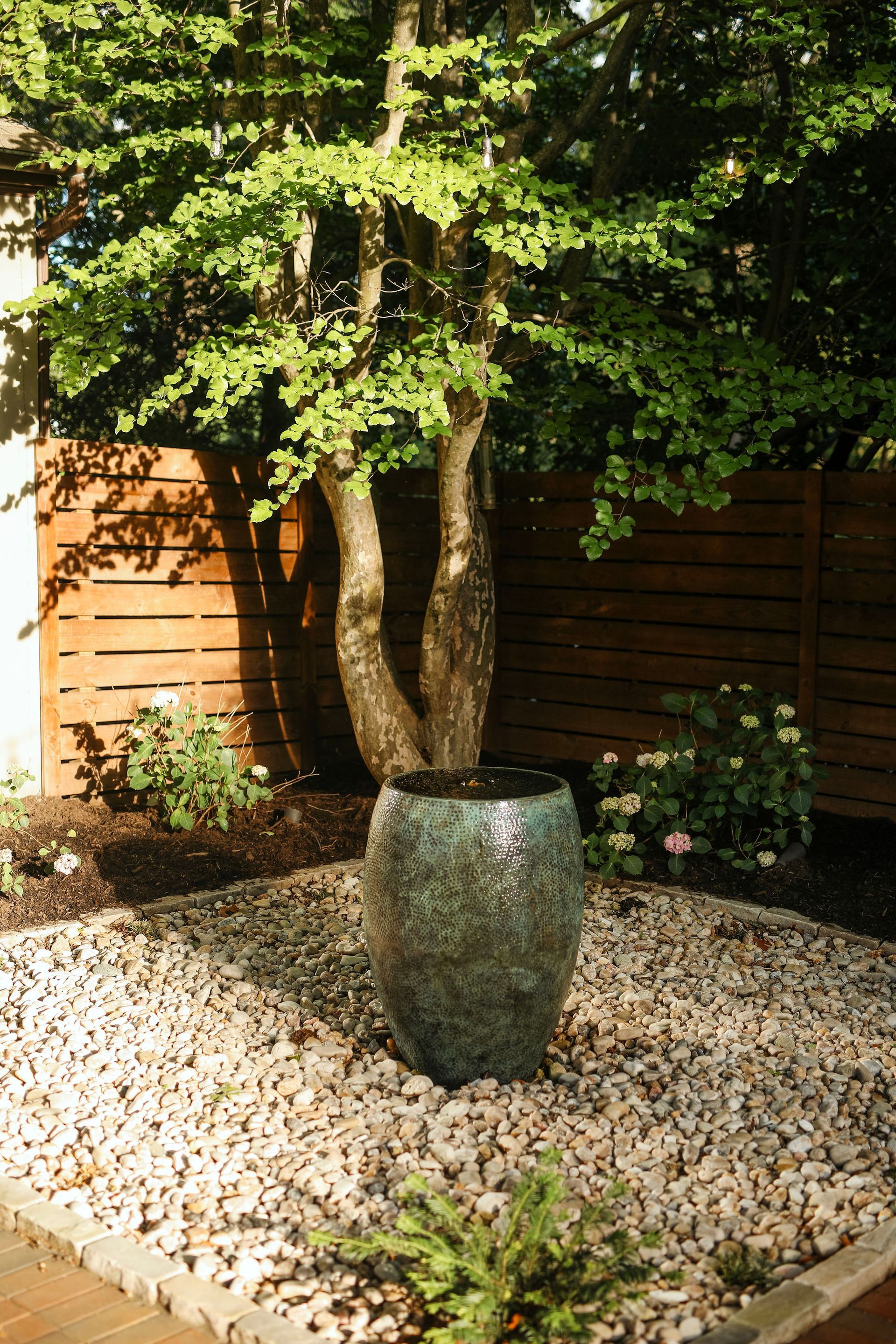 A turquoise glazed pot with a tree trunk is set in a bed of pebbles. A wood fence and greenery are in the background.