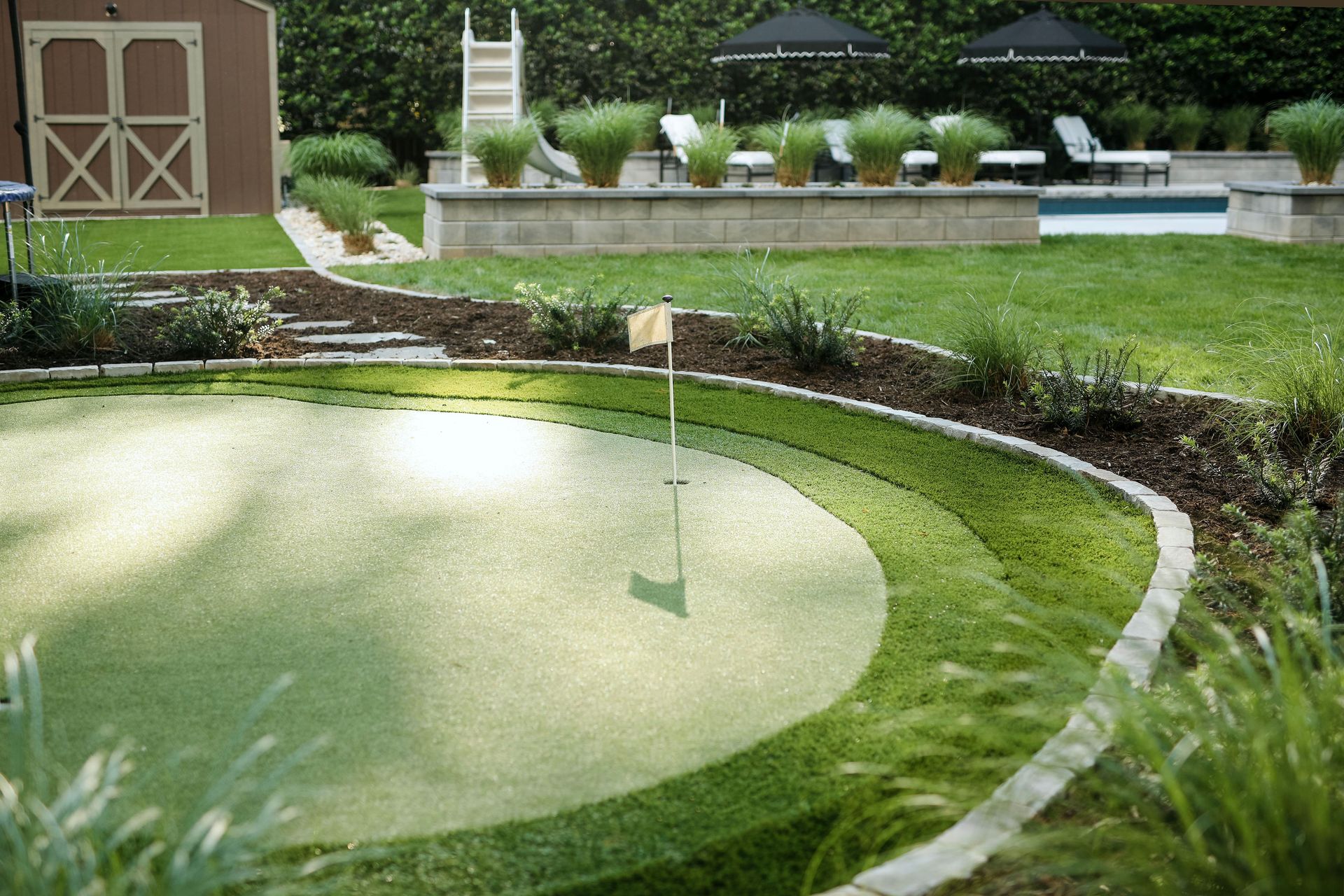 Backyard with a putting green, bordered by a curved stone wall.  A shed and pool are in the background, alongside grass and plants.