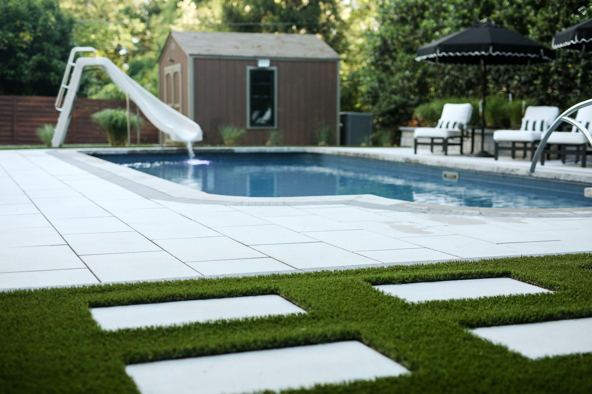 Pool area with a pool, slide, and lounge chairs. White pavers interspersed with turf create the ground, with a shed in the background.