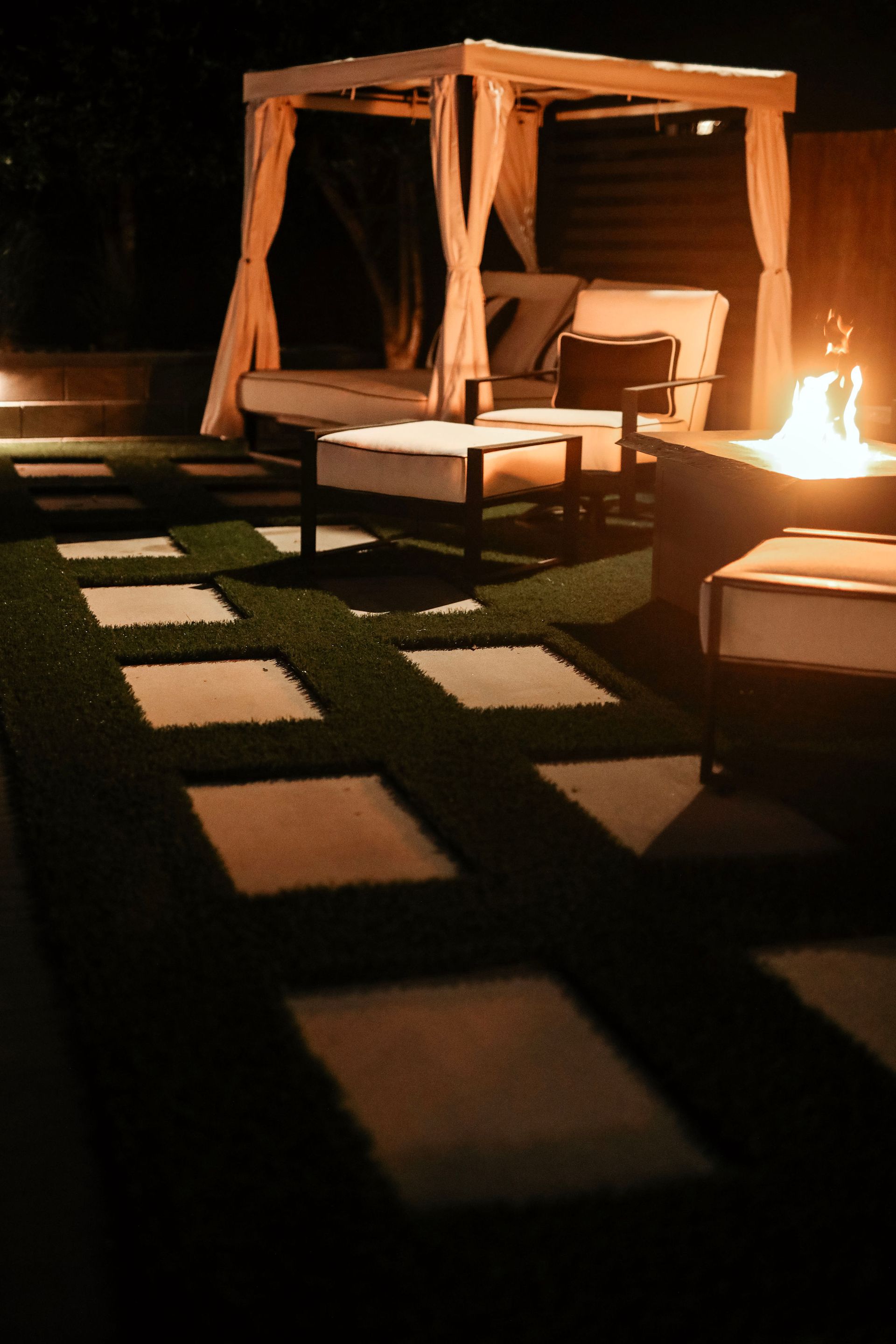 Night scene of a backyard patio with a seating area, a fire pit, and a pathway of stepping stones. Warm lighting creates a cozy atmosphere.