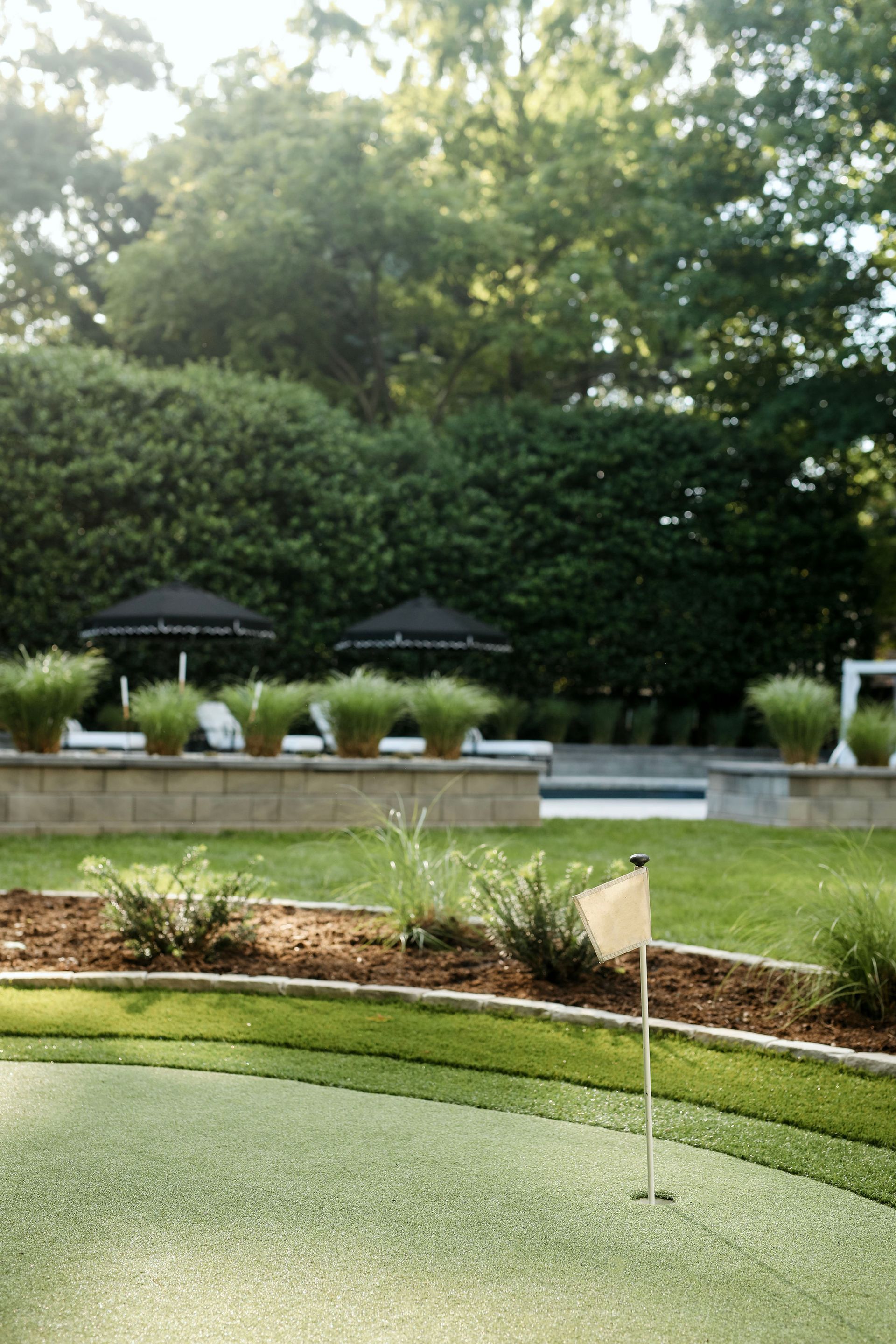 Putting green with a flag, overlooking a manicured lawn and a pool area with black umbrellas and green hedges.