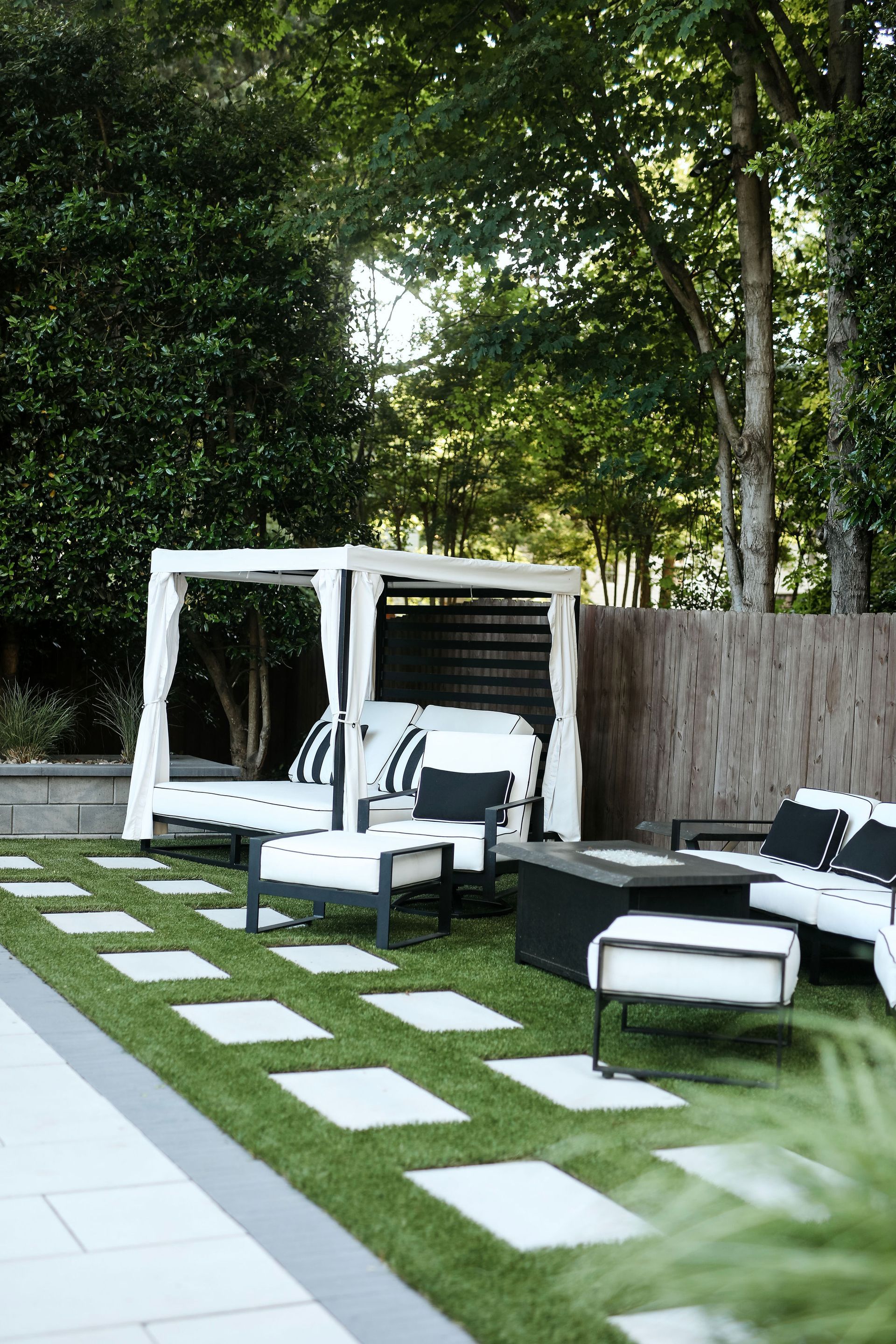 Outdoor seating area with white canopy bed, chairs, and a black fire pit on a lawn with square stone stepping stones.