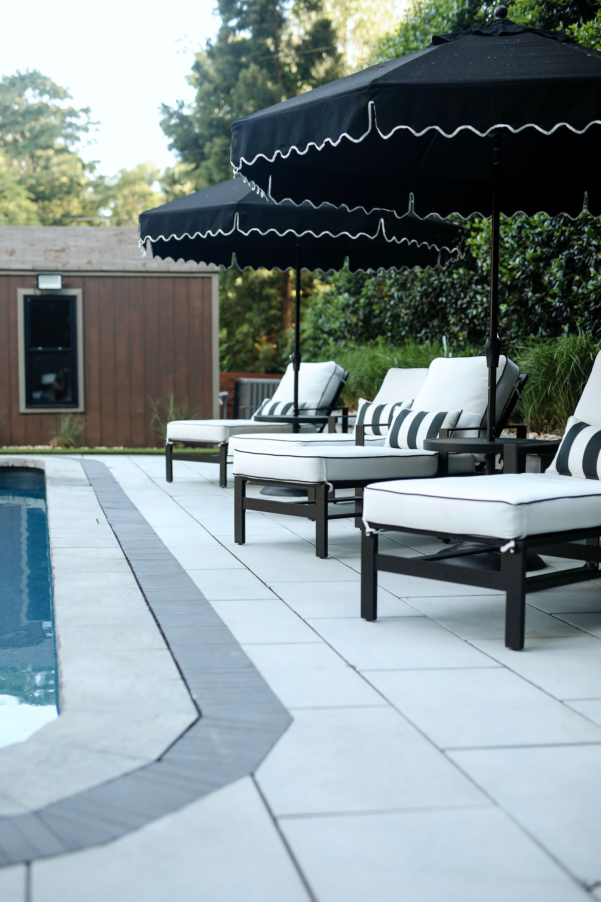 Poolside scene with black and white striped lounge chairs under black umbrellas with white trim. A wood-paneled building is in the background.
