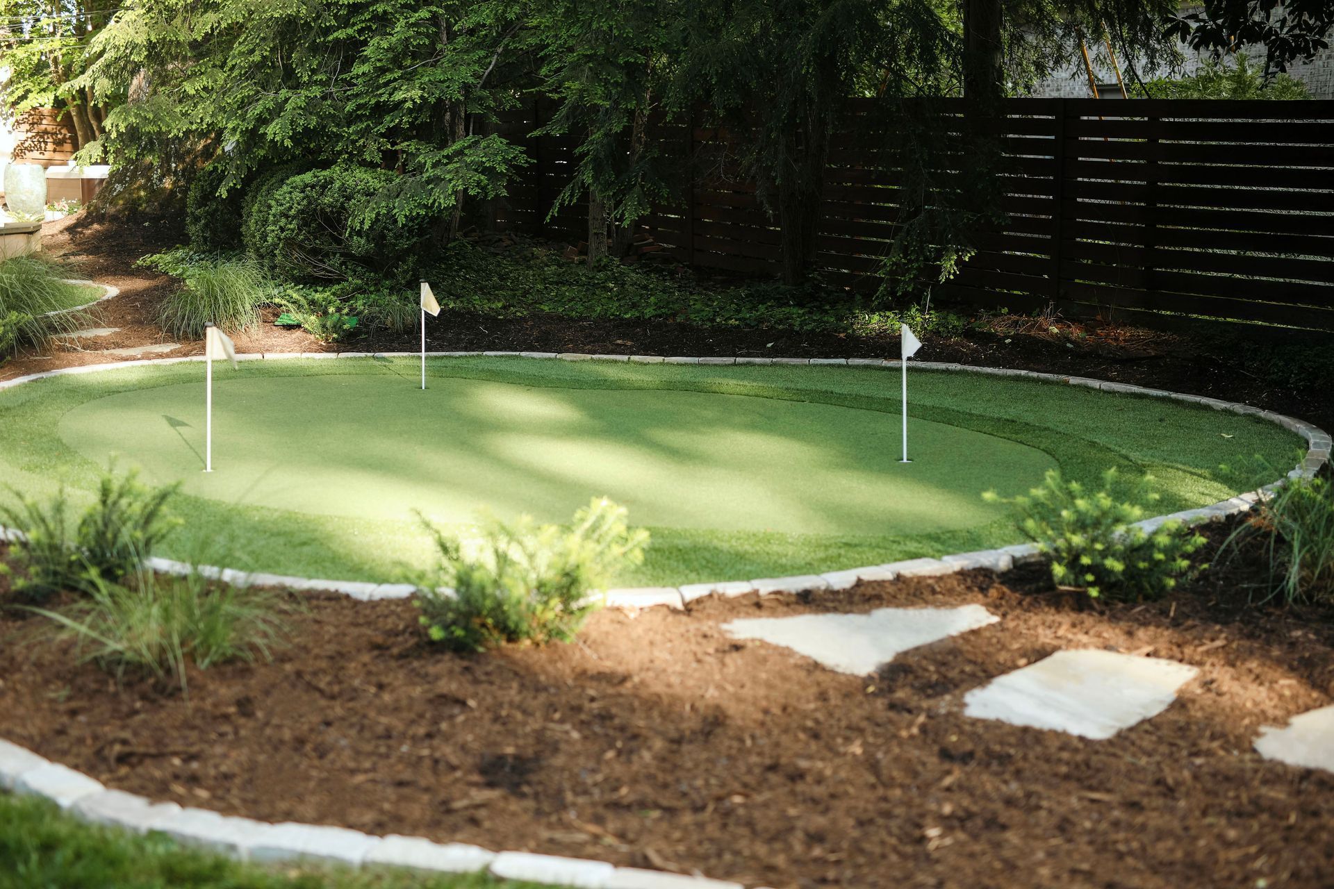 A backyard putting green with two flagsticks surrounded by mulch, stone edging, and landscaping.