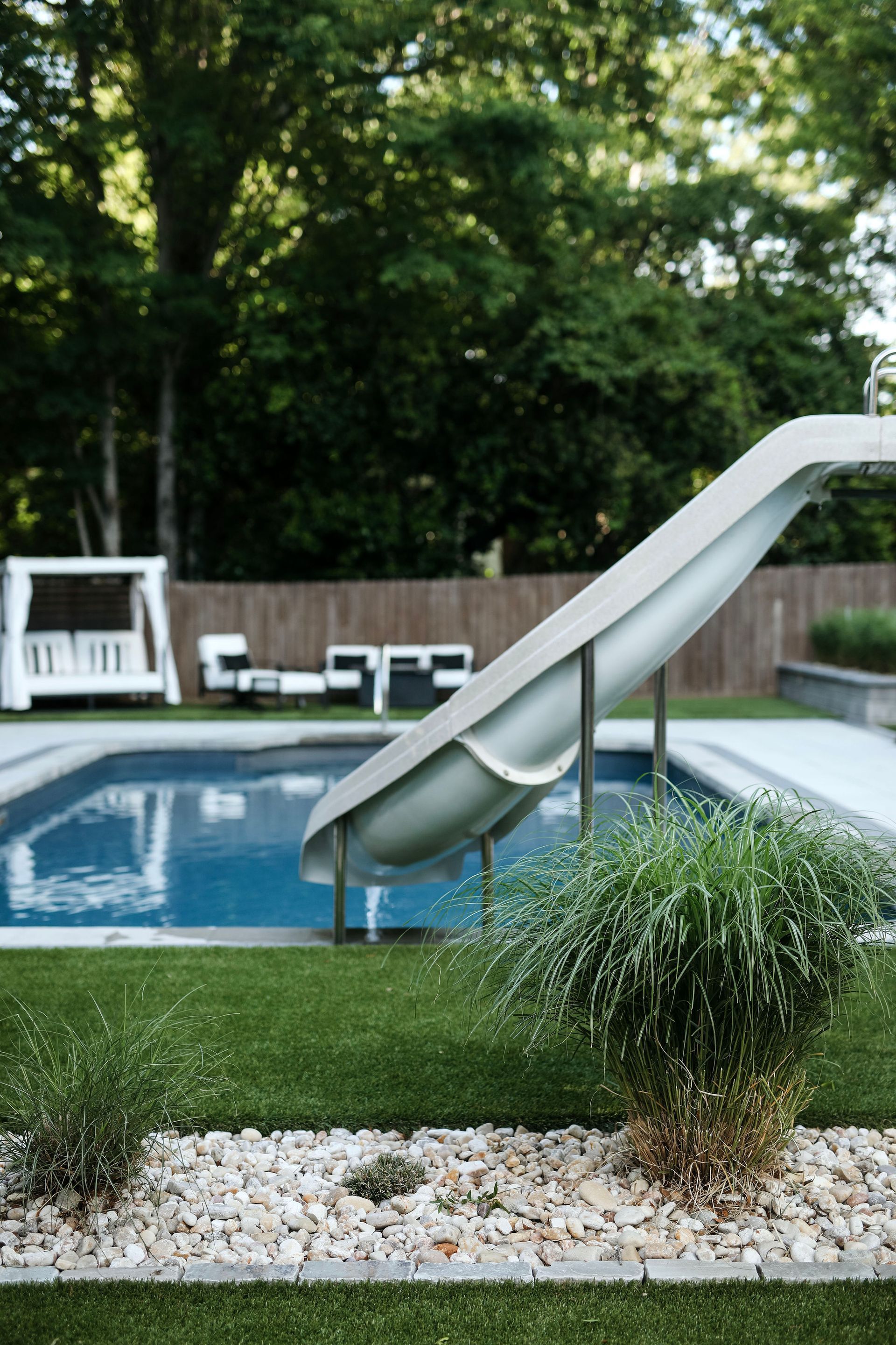 A backyard pool with a white water slide, surrounded by lush green grass and landscaping.
