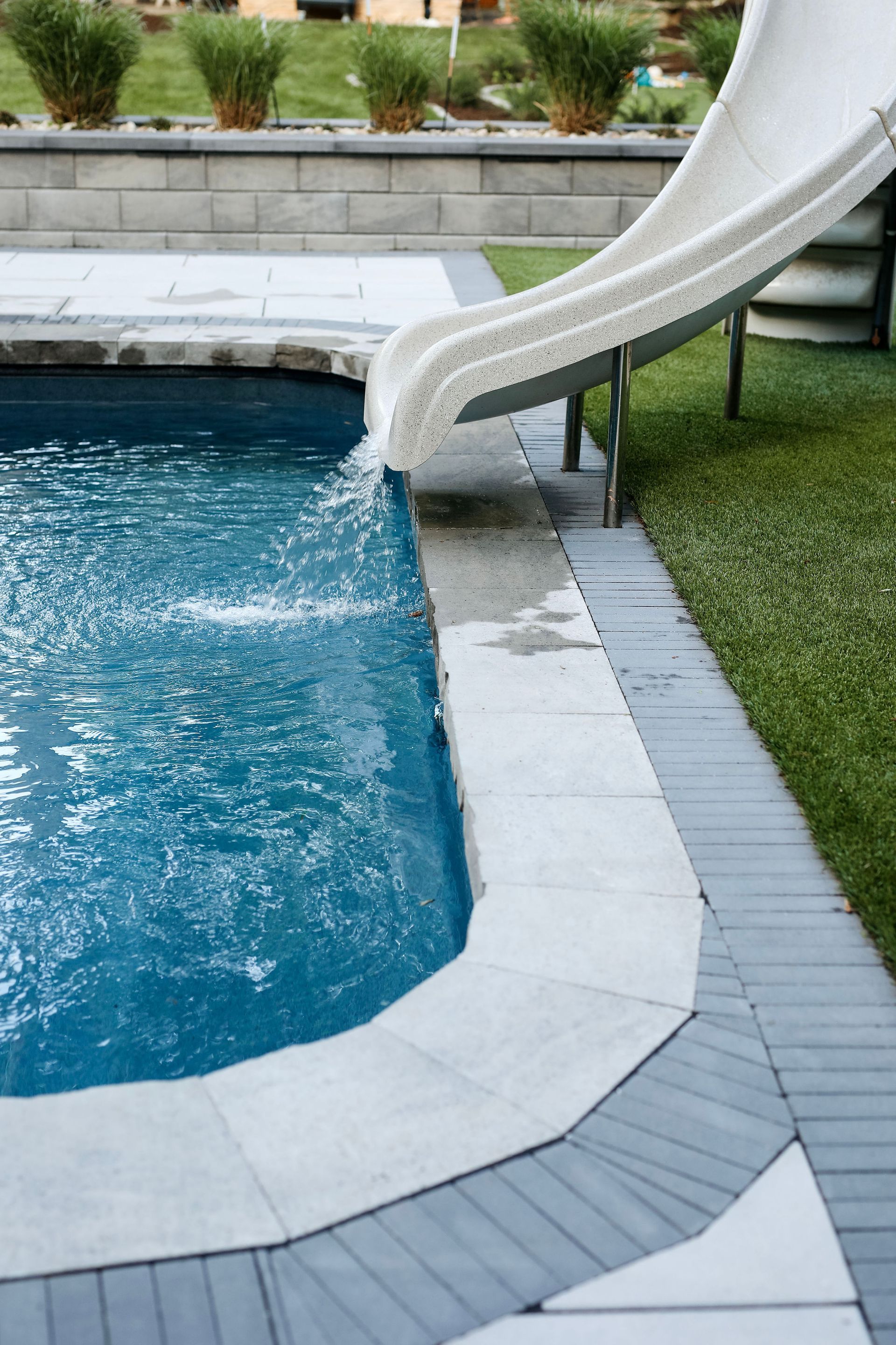 Pool with water slide; white slide descends into the blue water. Grey stone border surrounds the pool.