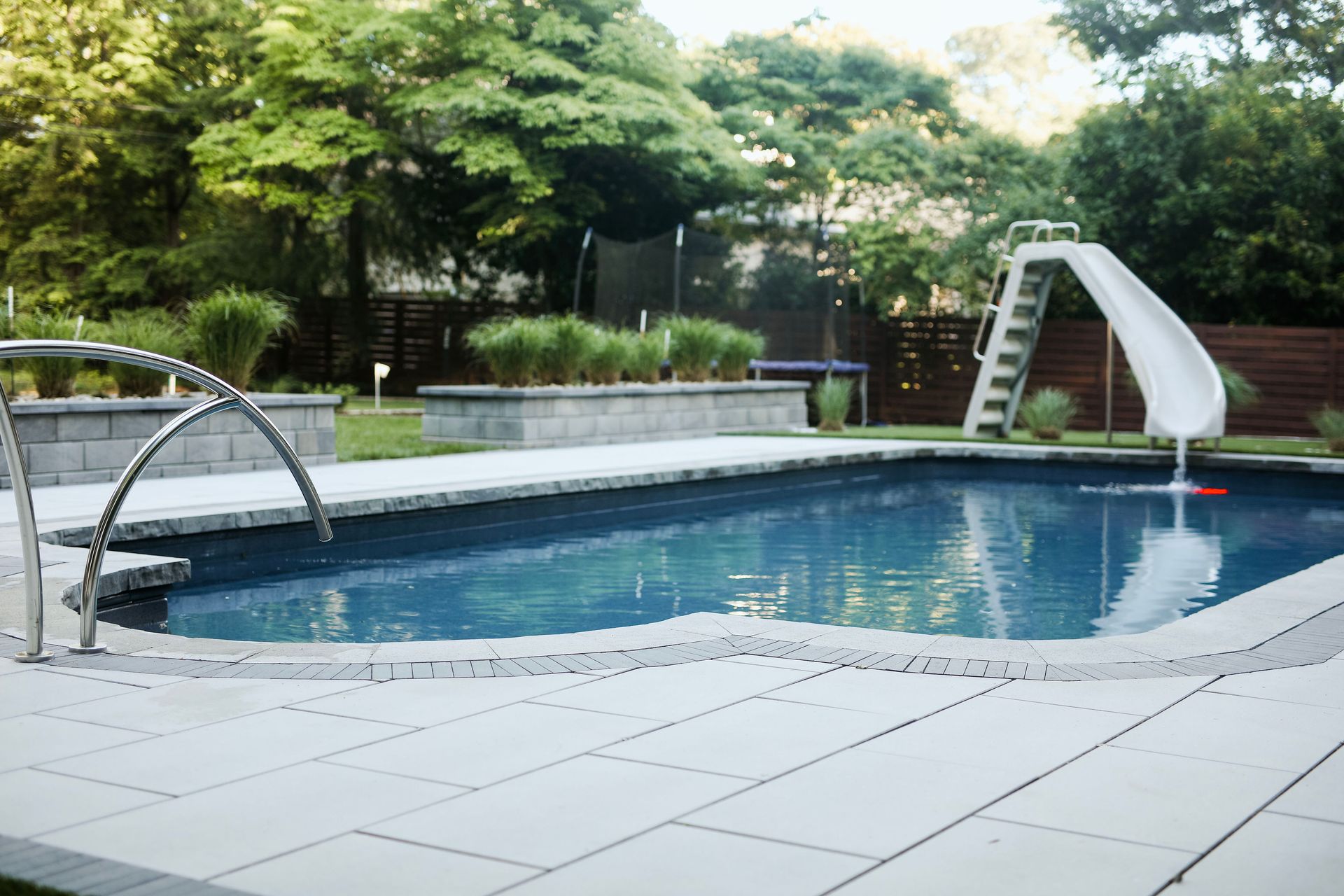 A swimming pool with a slide in a backyard. The pool has clear blue water, light-colored paving stones, and green trees in the background.