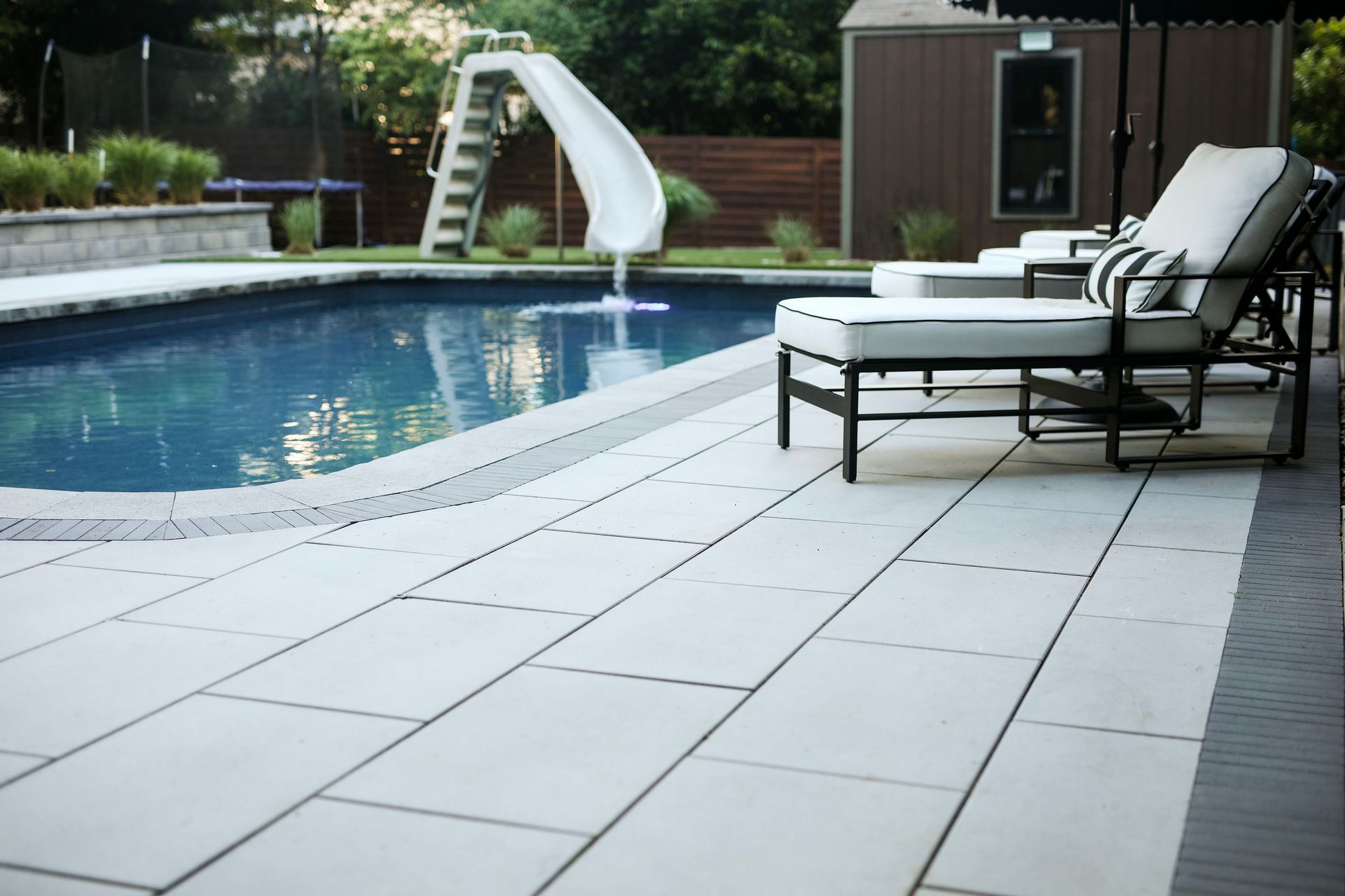Poolside scene with a pool, slide, lounge chairs, and light gray pavers. Lush green lawn and trees frame the background.