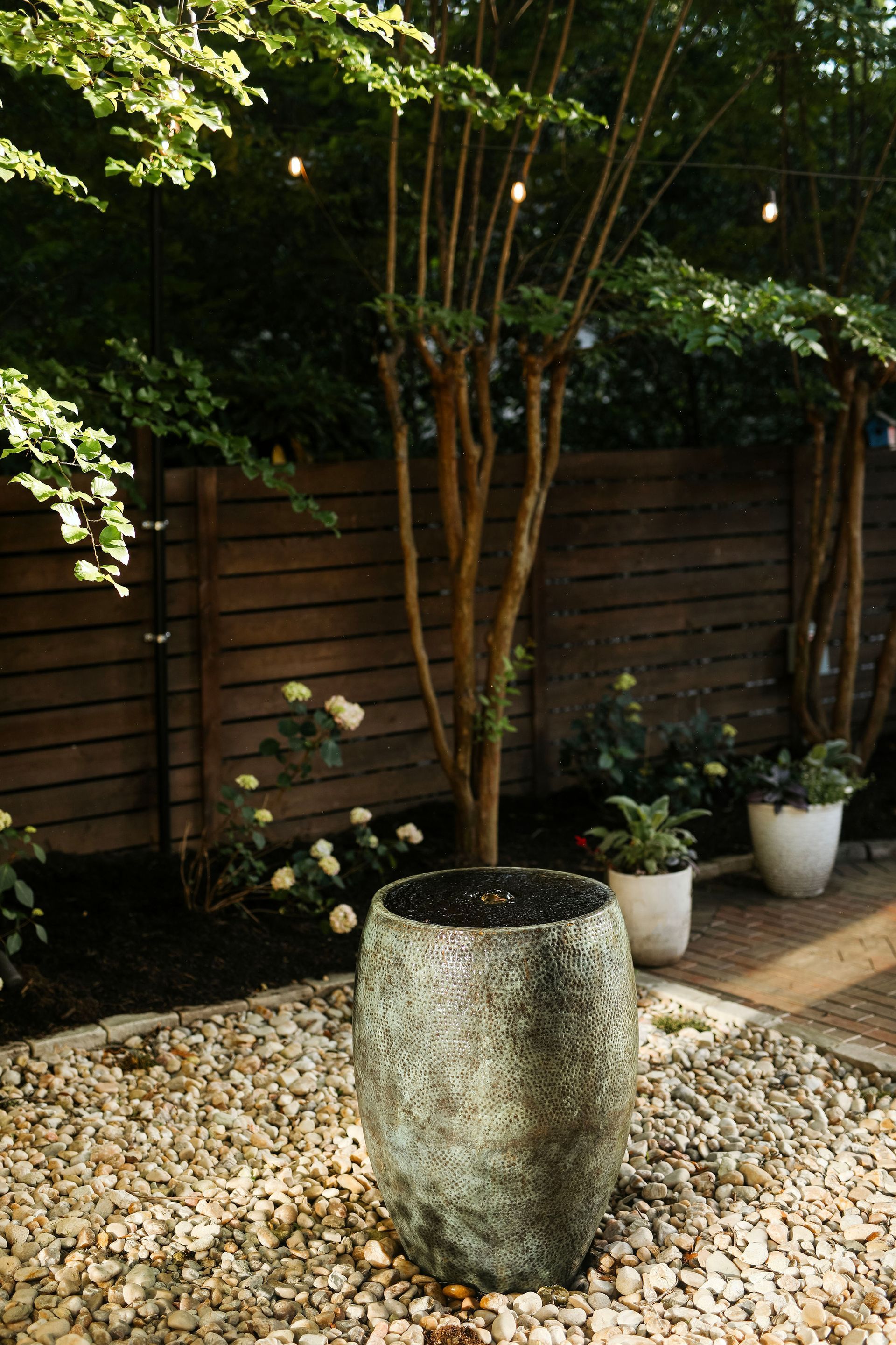 A garden scene featuring a textured, cylindrical pot on a bed of pebbles, with a wooden fence and trees in the background.