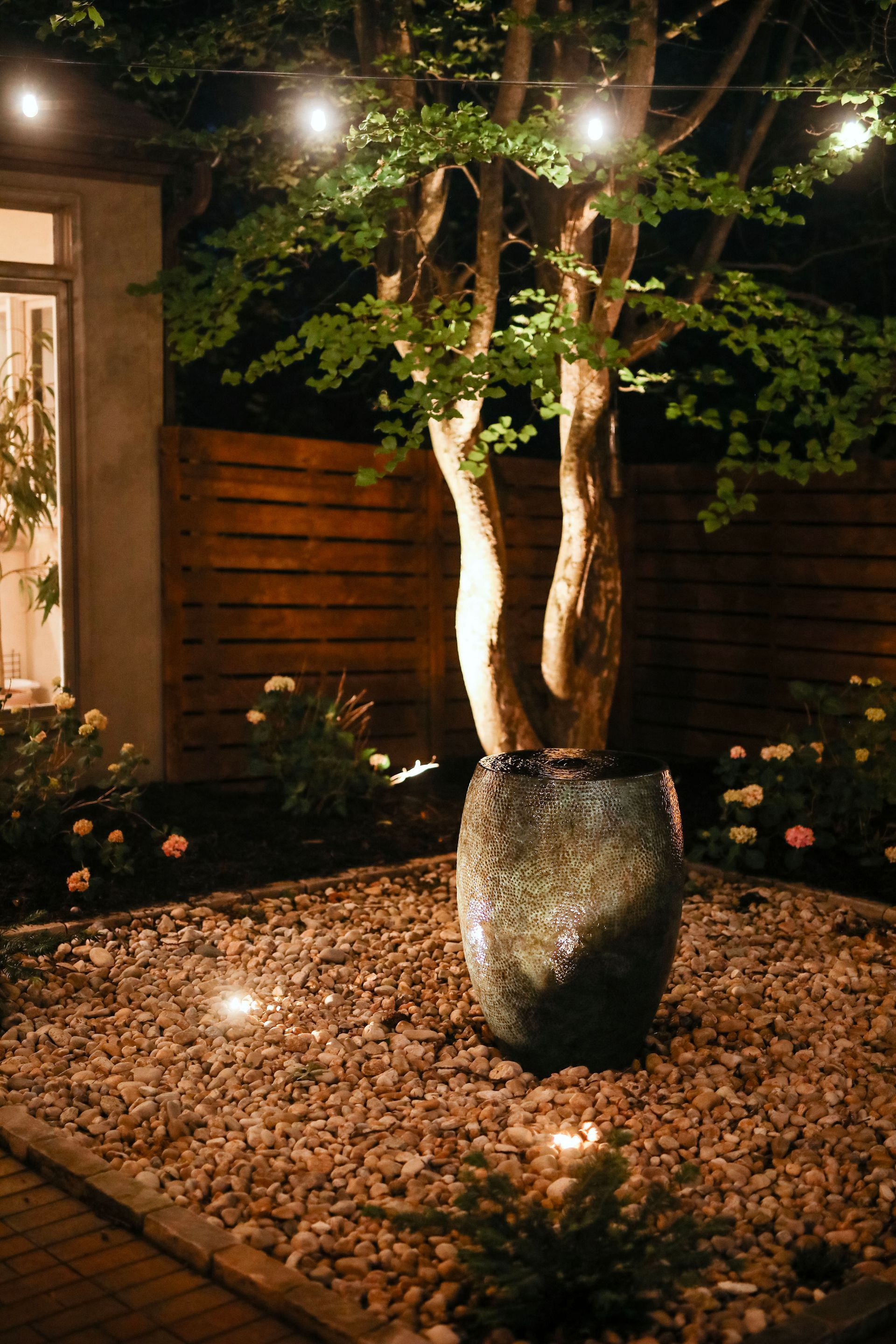 A nighttime backyard scene features a tree in a large glazed pot, lit by string lights and ground spotlights, surrounded by rocks.