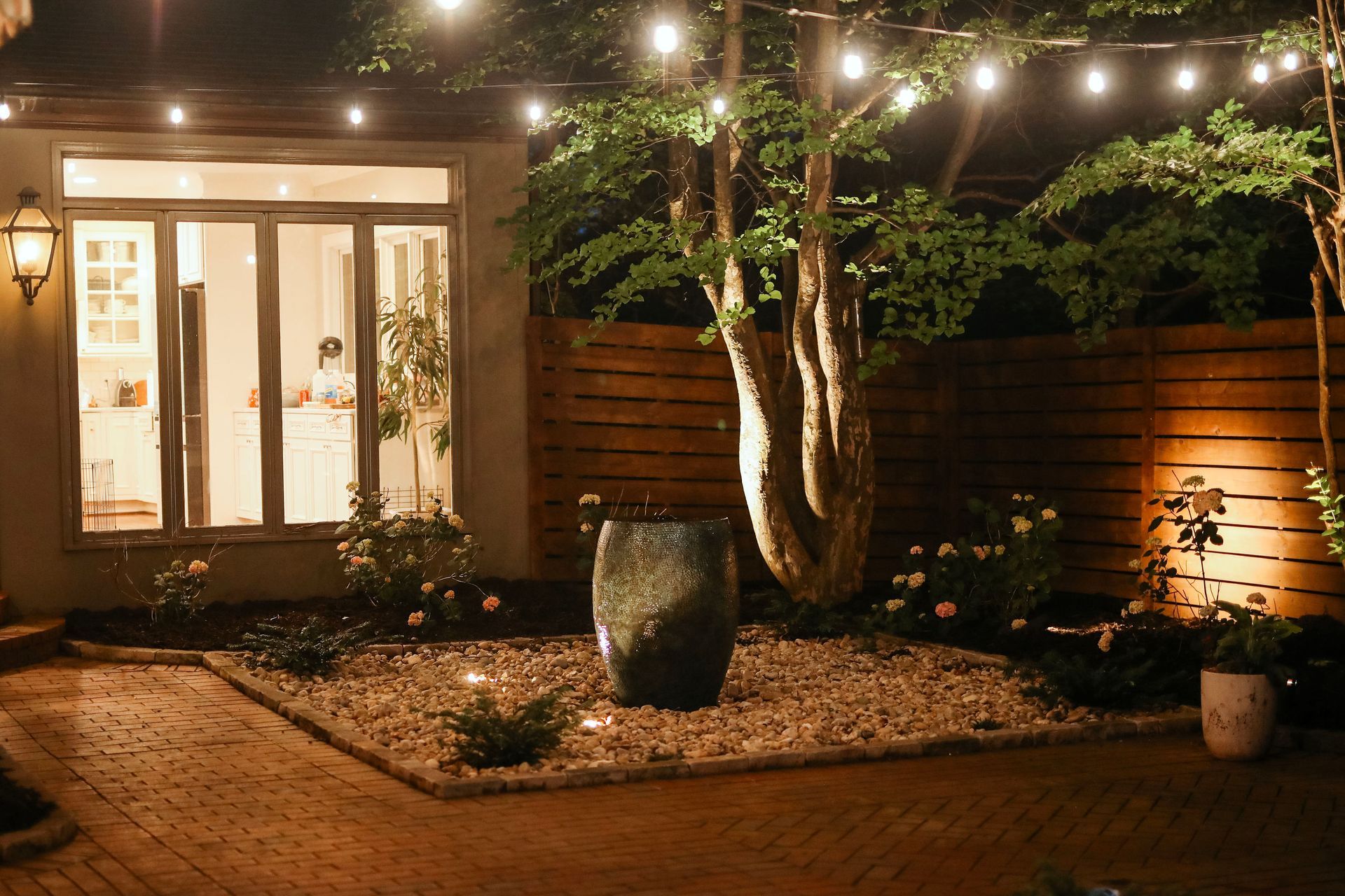 A cozy backyard patio at night, illuminated by string lights and spotlights. Features a tree, water fountain, and flowerbeds next to a house with glass doors.