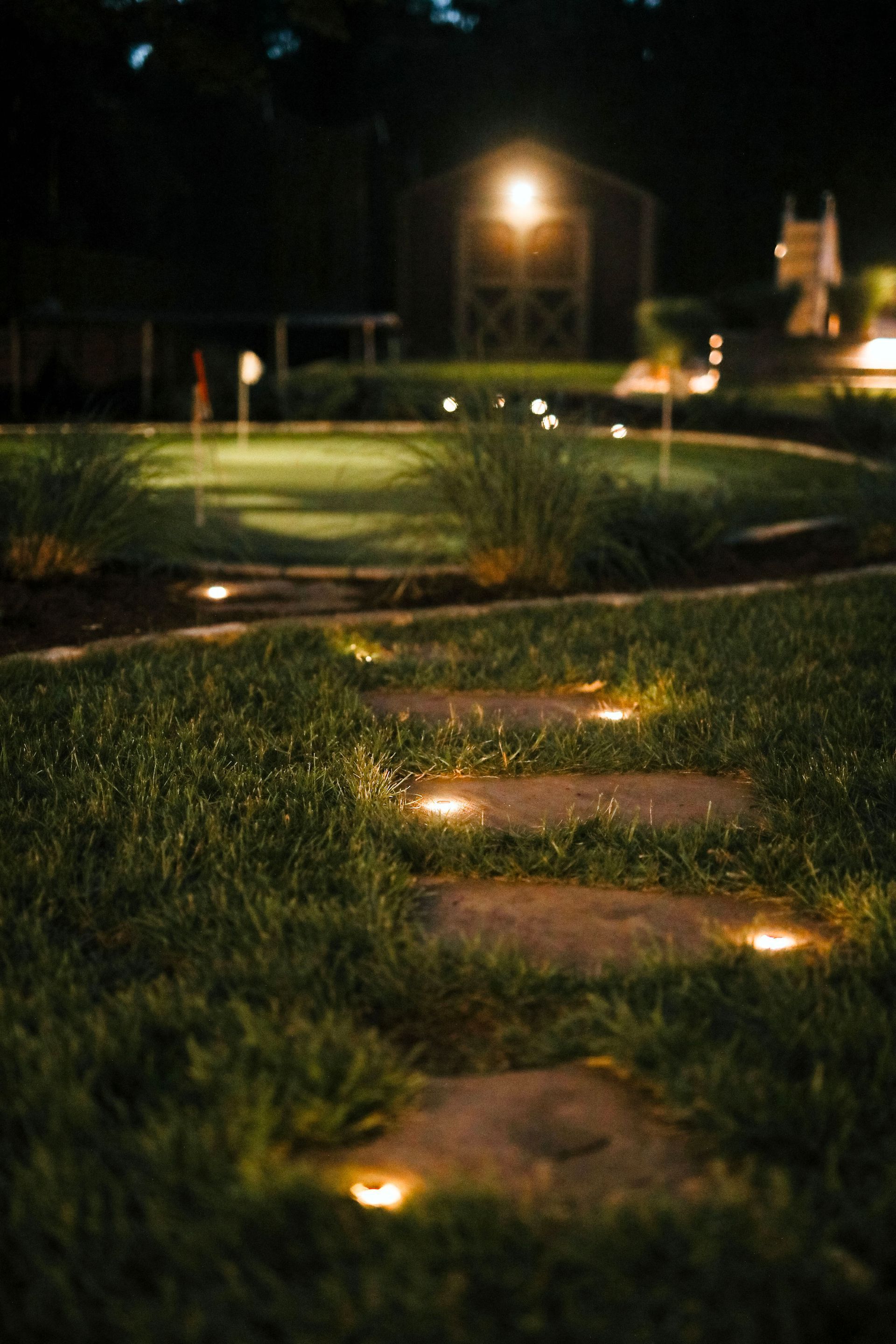 A stone path with glowing lights leads toward a lit-up barn and a putting green at night.