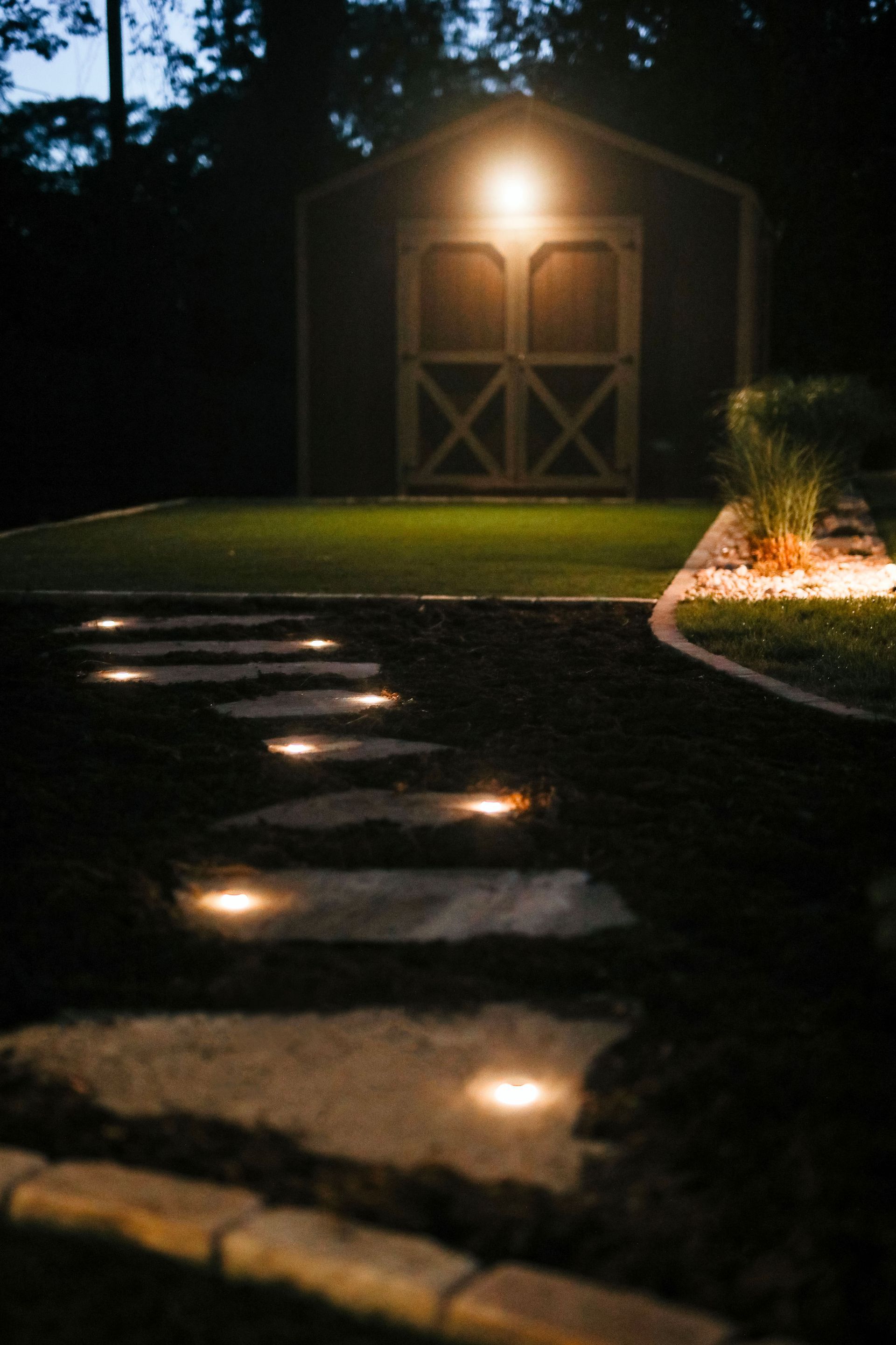 A stone path with embedded lights leads to a lit shed in a backyard at night.