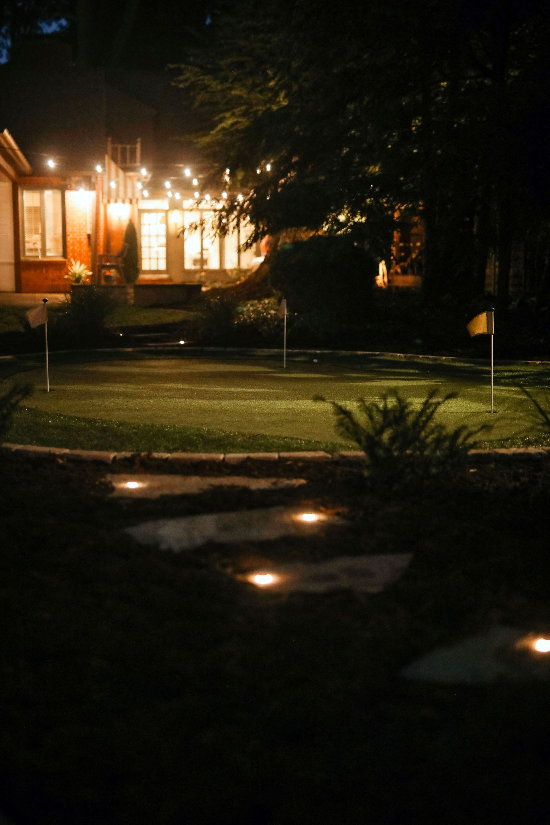 Nighttime backyard scene with a putting green, illuminated stone pathway, and house in the background strung with lights.