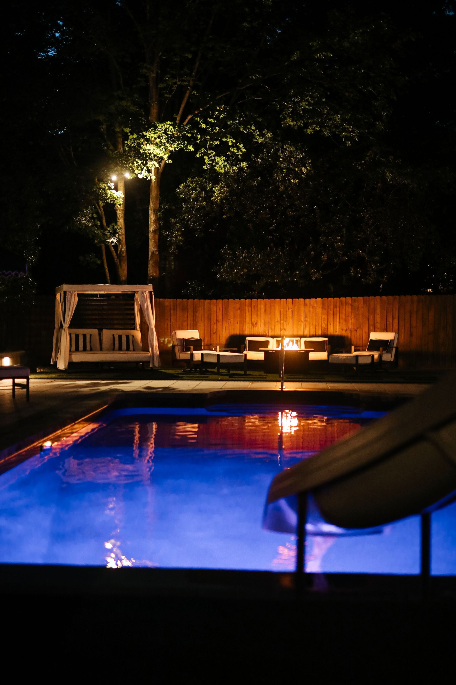 Nighttime outdoor pool scene with string lights, lounge chairs, and a cabana; the water glows blue.