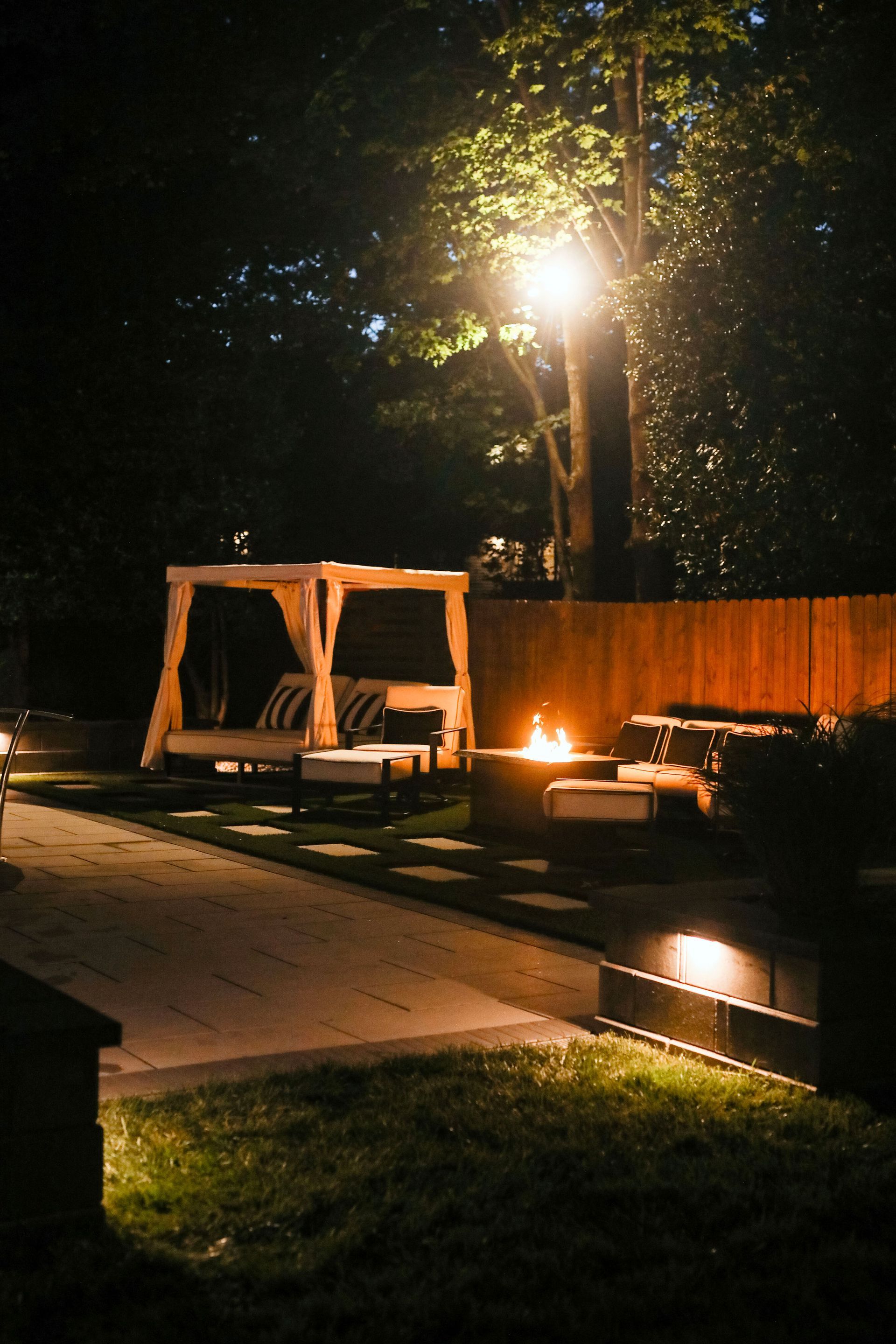 Night scene of an outdoor lounge with a lit fire pit and canopy bed. Warm light illuminates the wooden structures and surrounding foliage.
