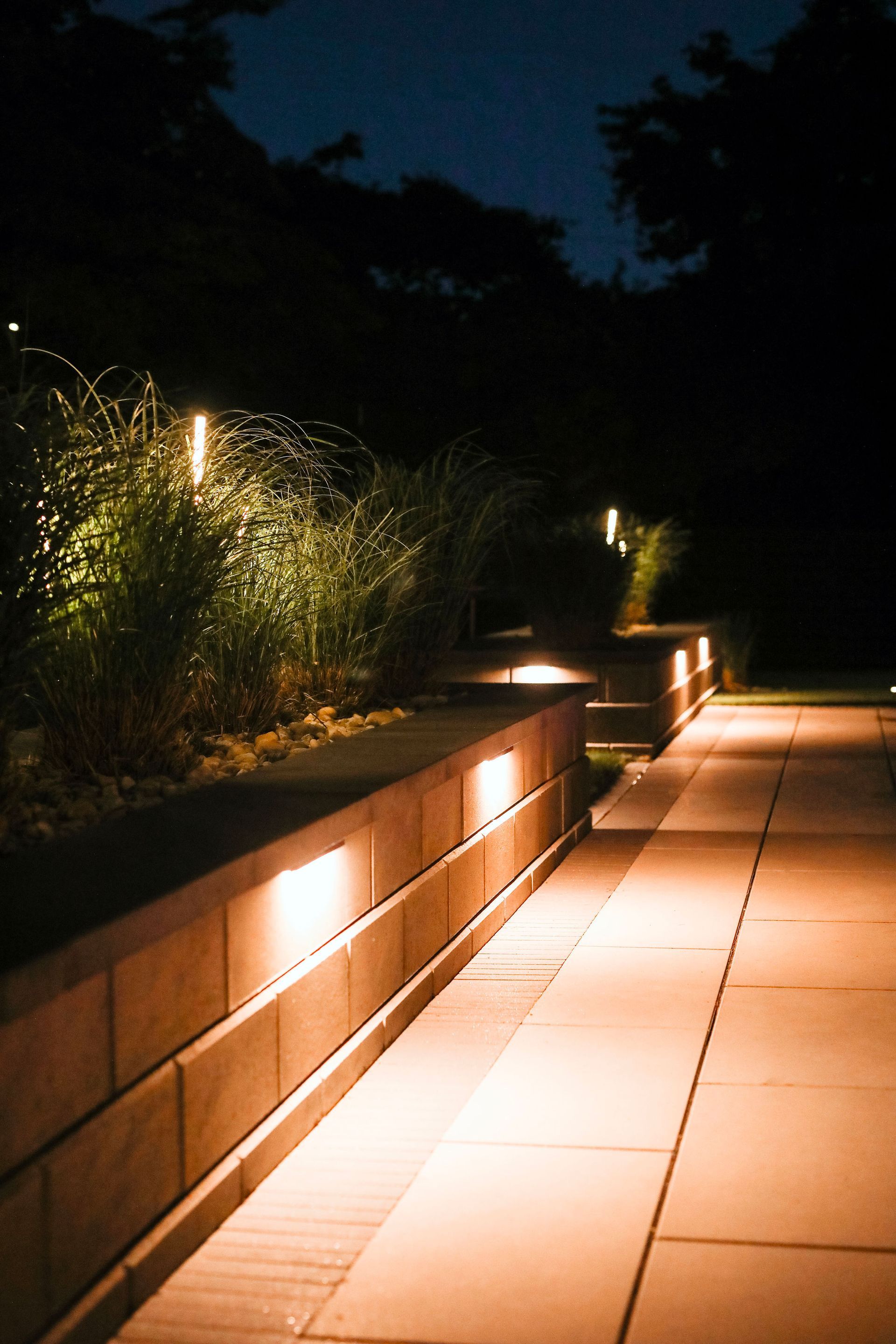 Lit pathway and retaining wall at night. Warm lights illuminate the path and the textured brick wall with lush greenery.