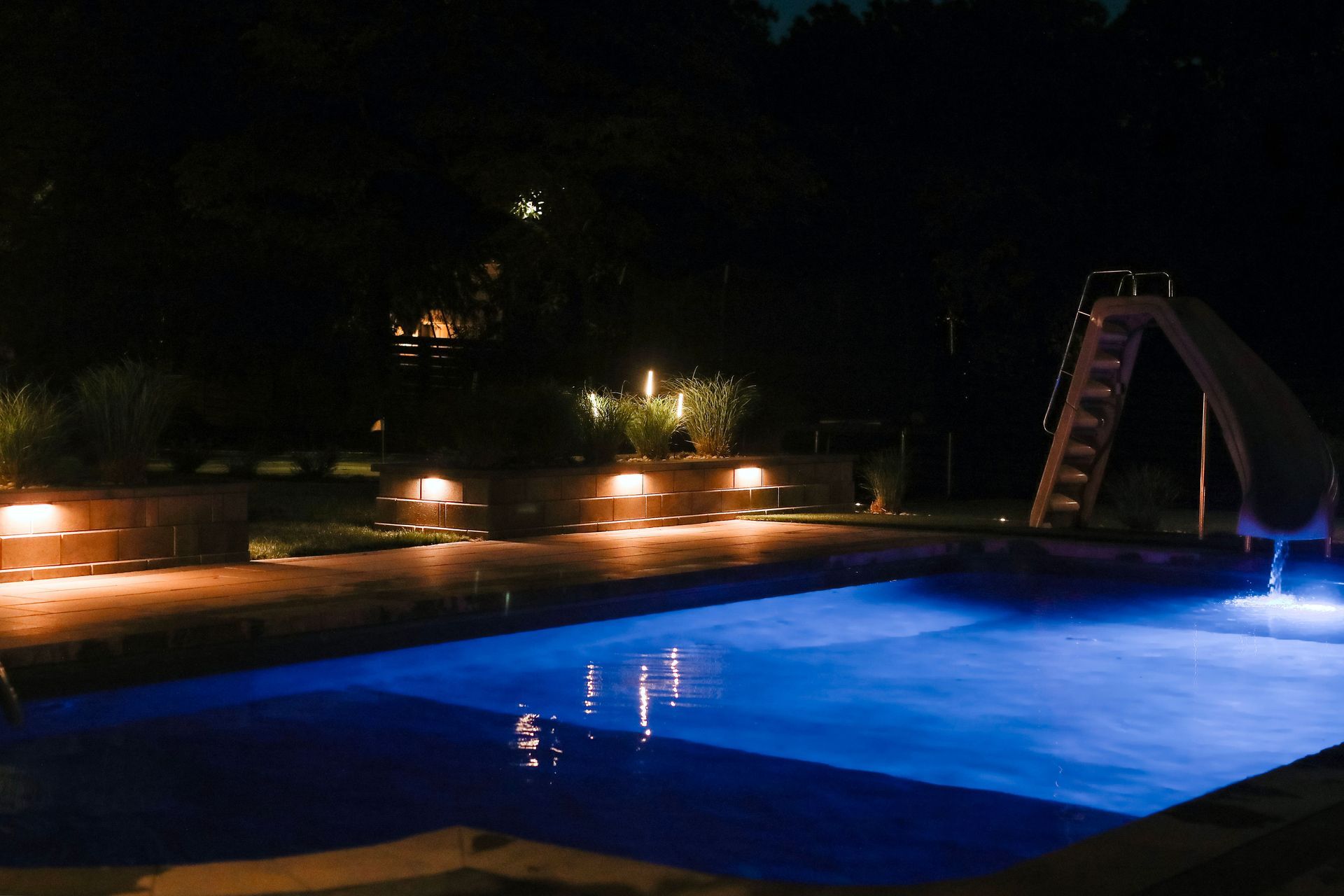 Nighttime pool with blue water, slide, and surrounding landscape lighting illuminating the pool area.