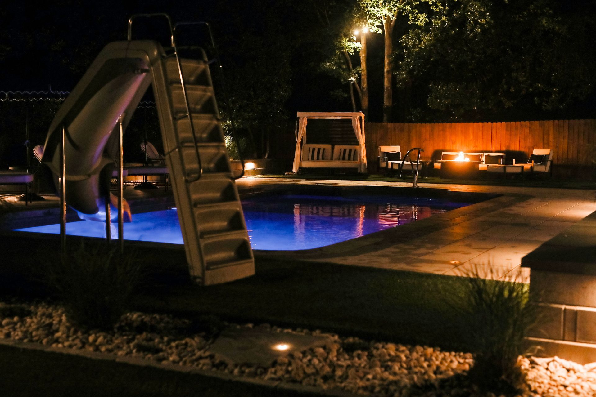 Nighttime shot of a pool with a slide, illuminated by blue underwater lights and ambient lighting. Wooden seating and a swing bed are nearby.