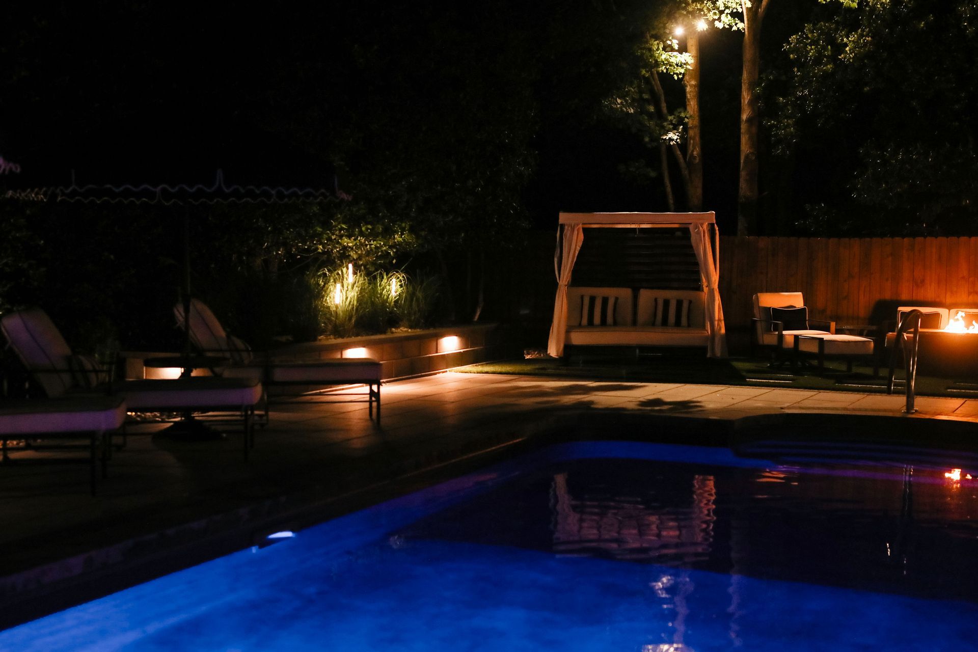 Nighttime backyard scene with a pool reflecting blue light. Lounge chairs, a canopy bed, and a fire pit are lit by warm lights.