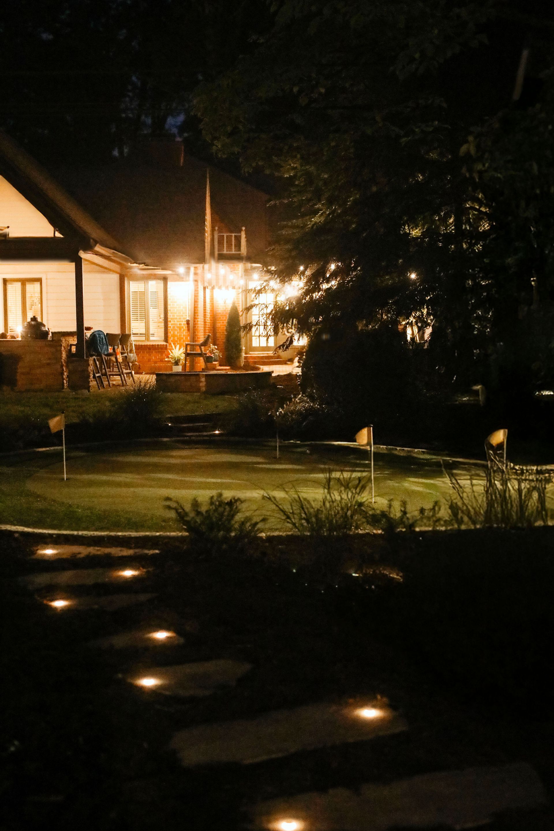 Pathway lit by ground lights leading to a house at night. Warm light shines from the house windows and yard.