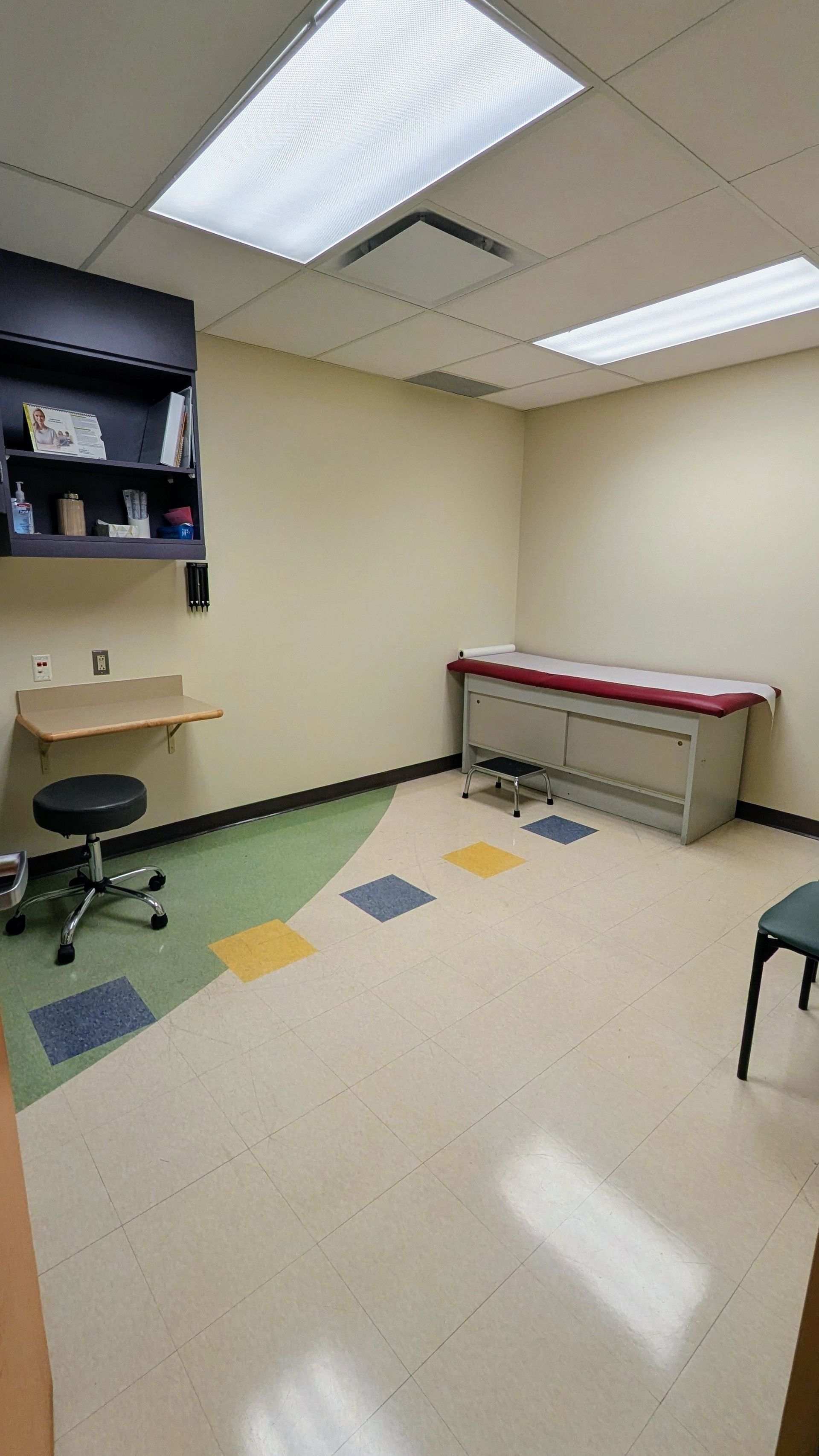 Small beige exam room with cabinets, exam table, stool, and tiled floor under fluorescent lights.