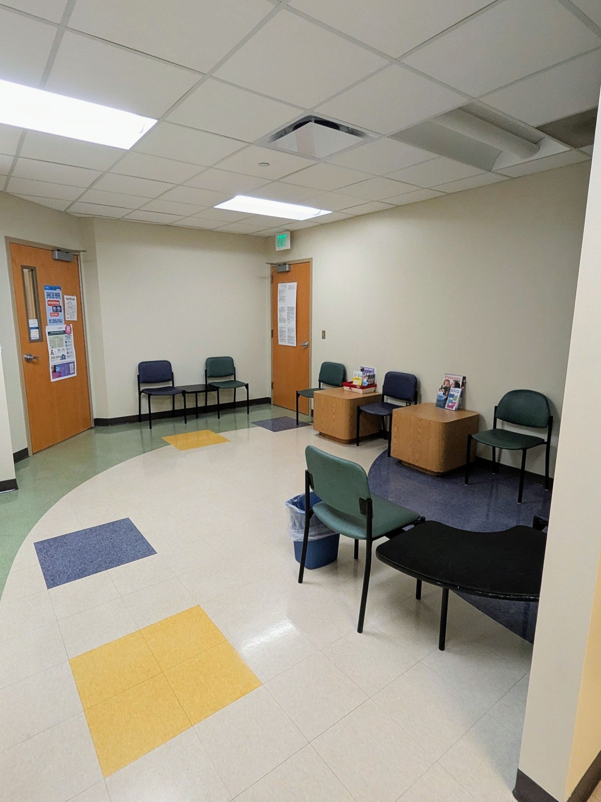 Empty waiting room with chairs, tables, colorful floor tiles, and doors along beige walls.
