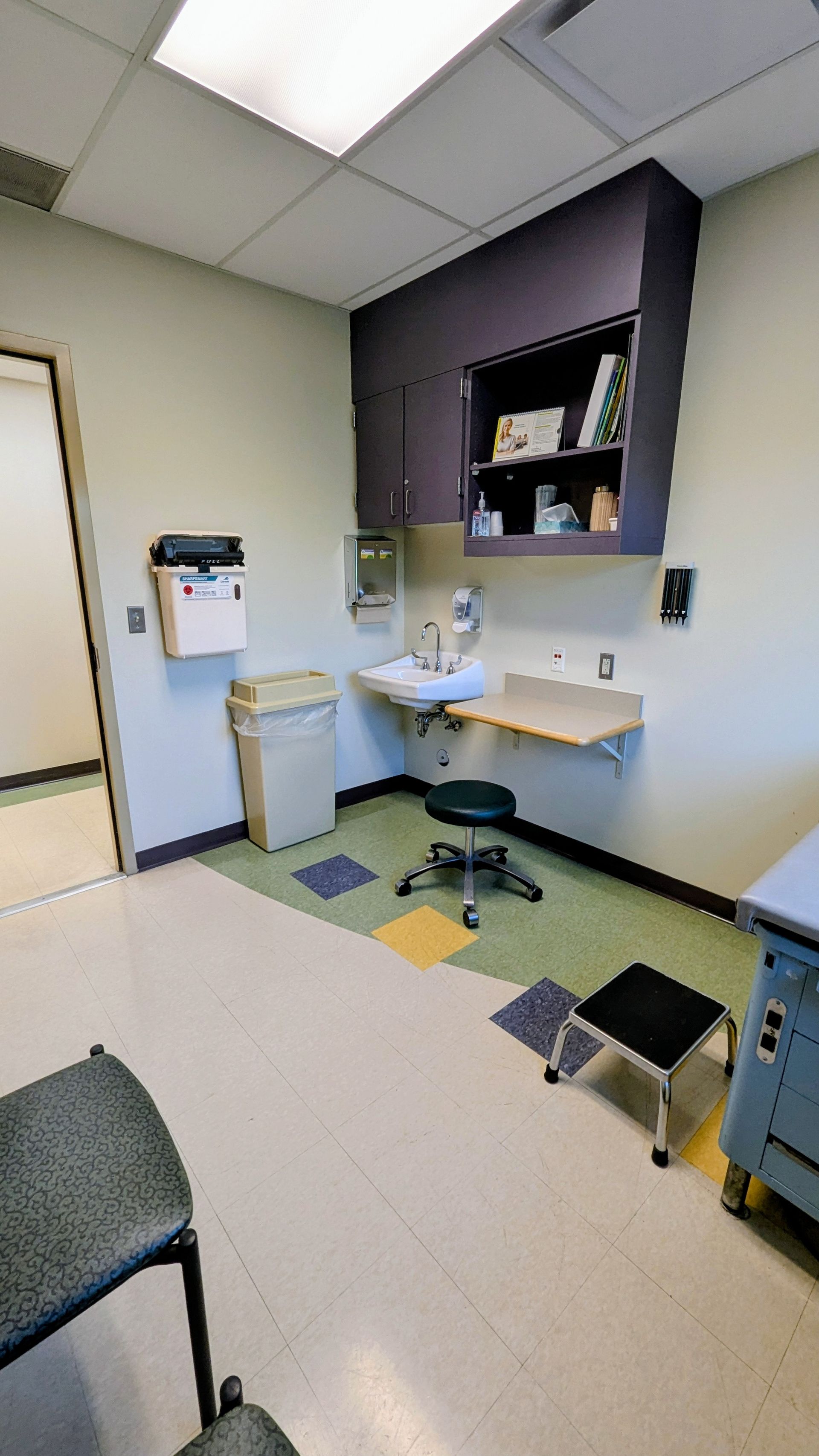 Small medical exam room with a sink, counter, cabinets, and a stool under fluorescent lighting.