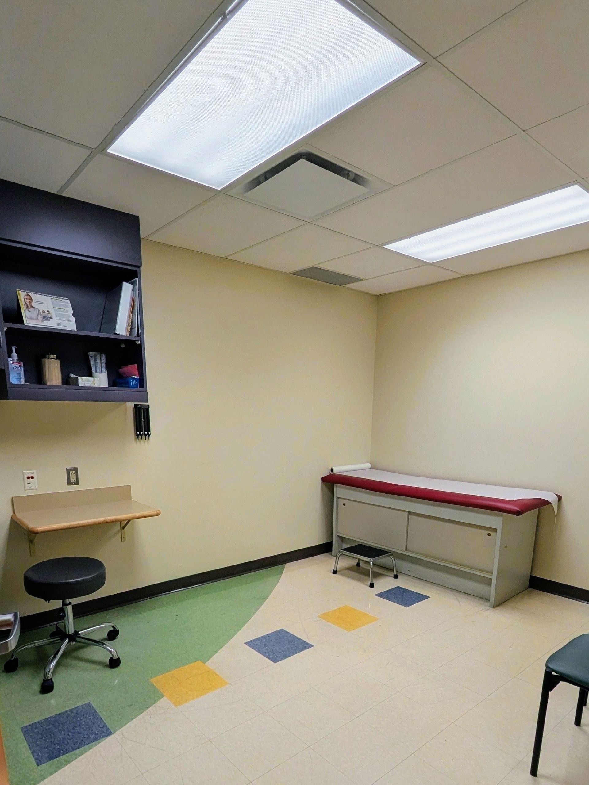 Small examination room with cabinets, stool, desk, examination table, and colorful floor tiles