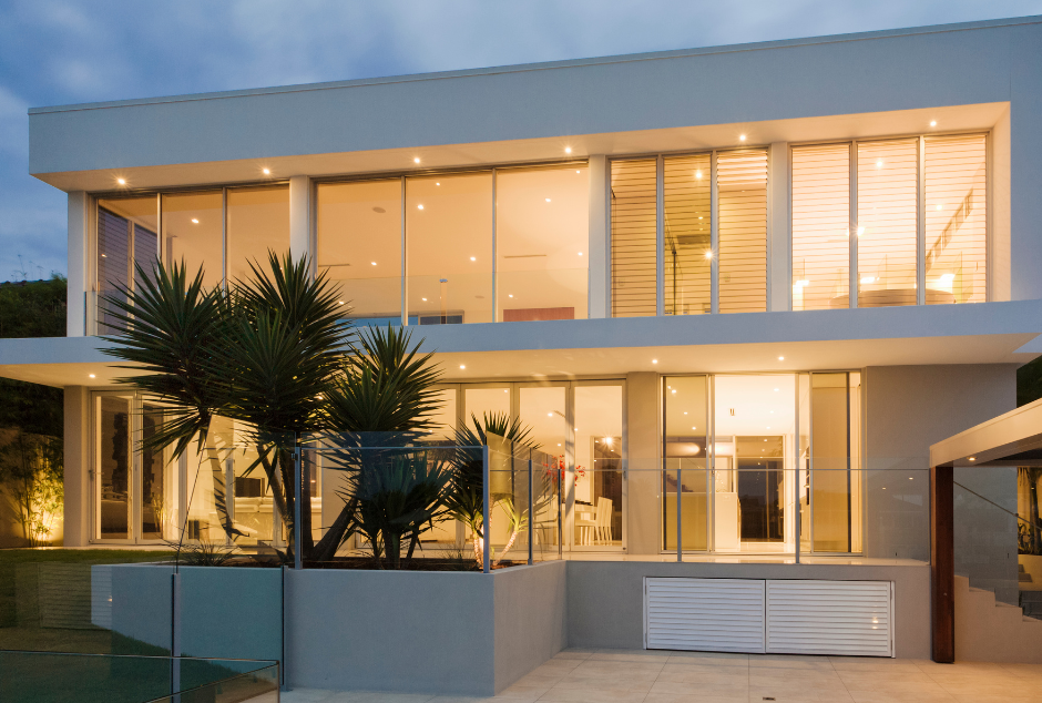 Modern white house with large windows, pool, and lush landscaping at dusk.