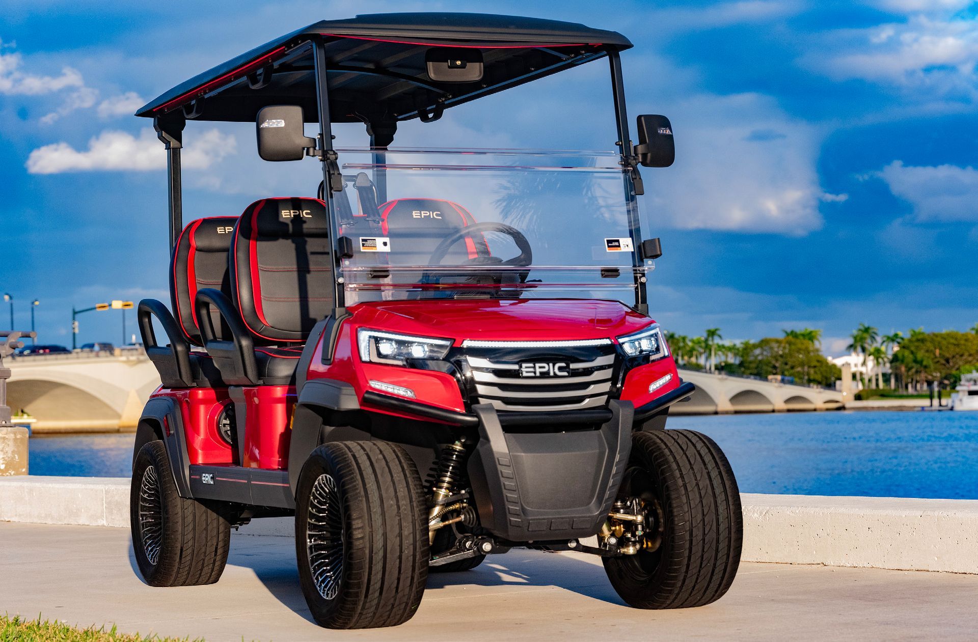 Red and black EPIC golf cart parked near water and a bridge, under a partly cloudy sky.