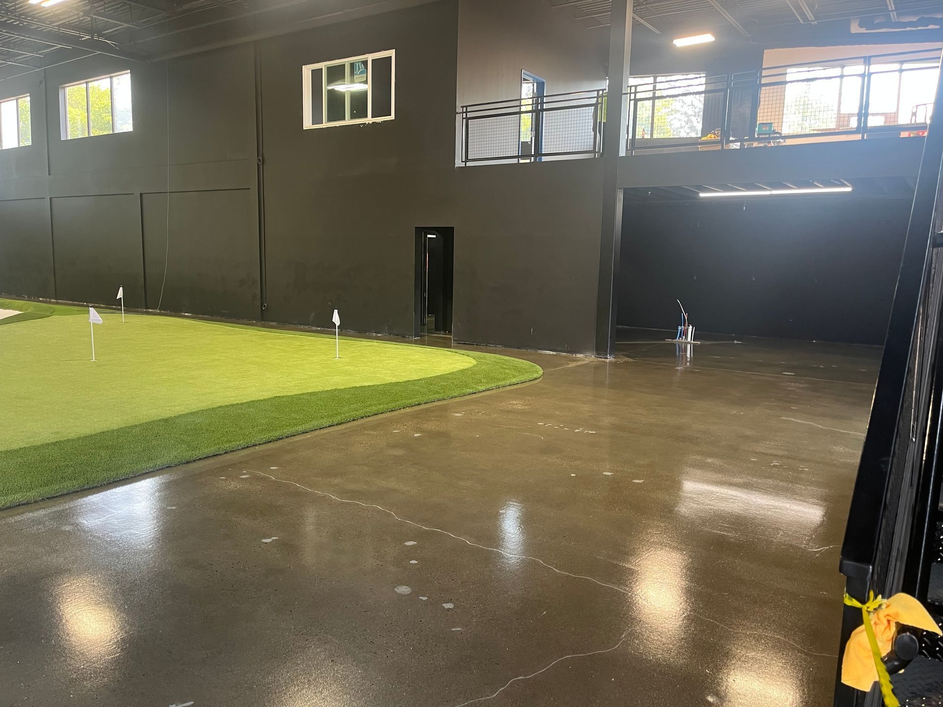 Indoor putting green next to wet concrete floor. Dark walls, upper level, with windows and black door.