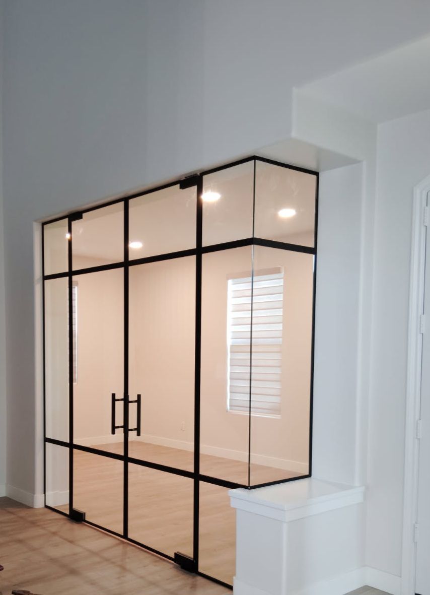 Glass-walled room with black metal framing and door handles. Interior with overhead lighting, white walls, and wood floor.